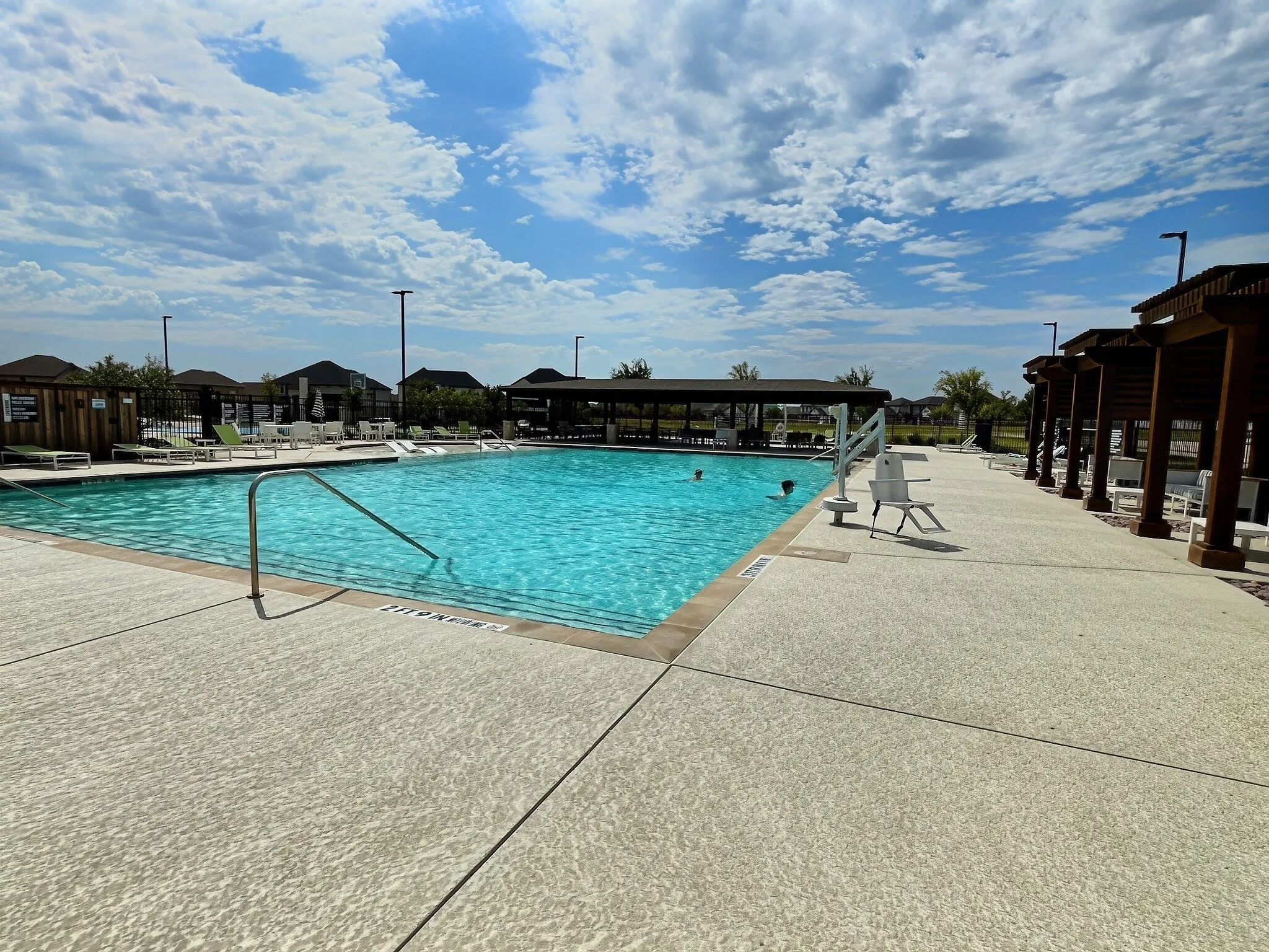 Expansive swimming pool with integrated access ramp, surrounded by a textured concrete deck, and shaded pergola structures
