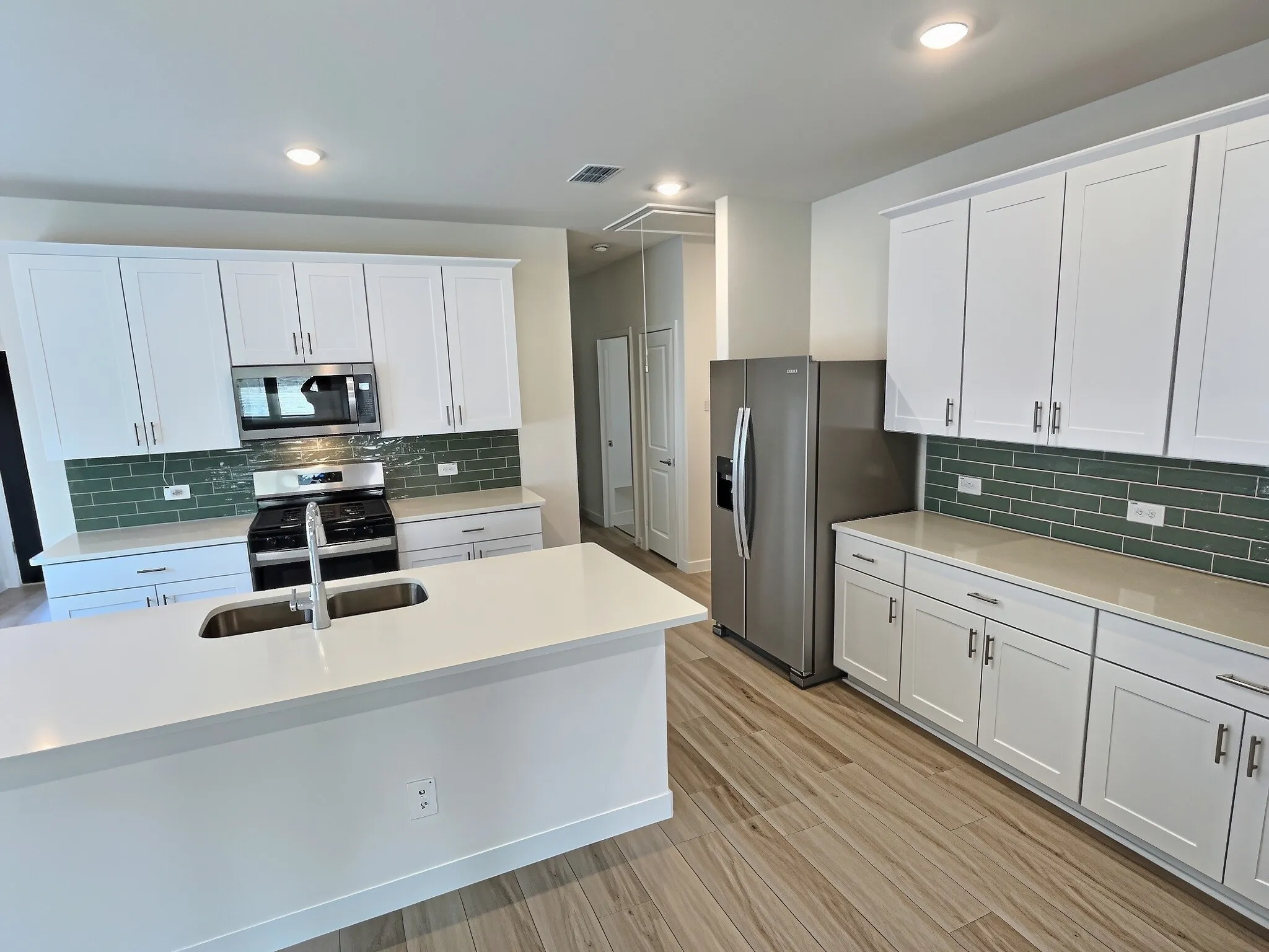 Kitchen featuring a center island with an undermount sink, white cabinetry, integrated stainless steel appliances, green subway tile backsplash, and light-toned countertops