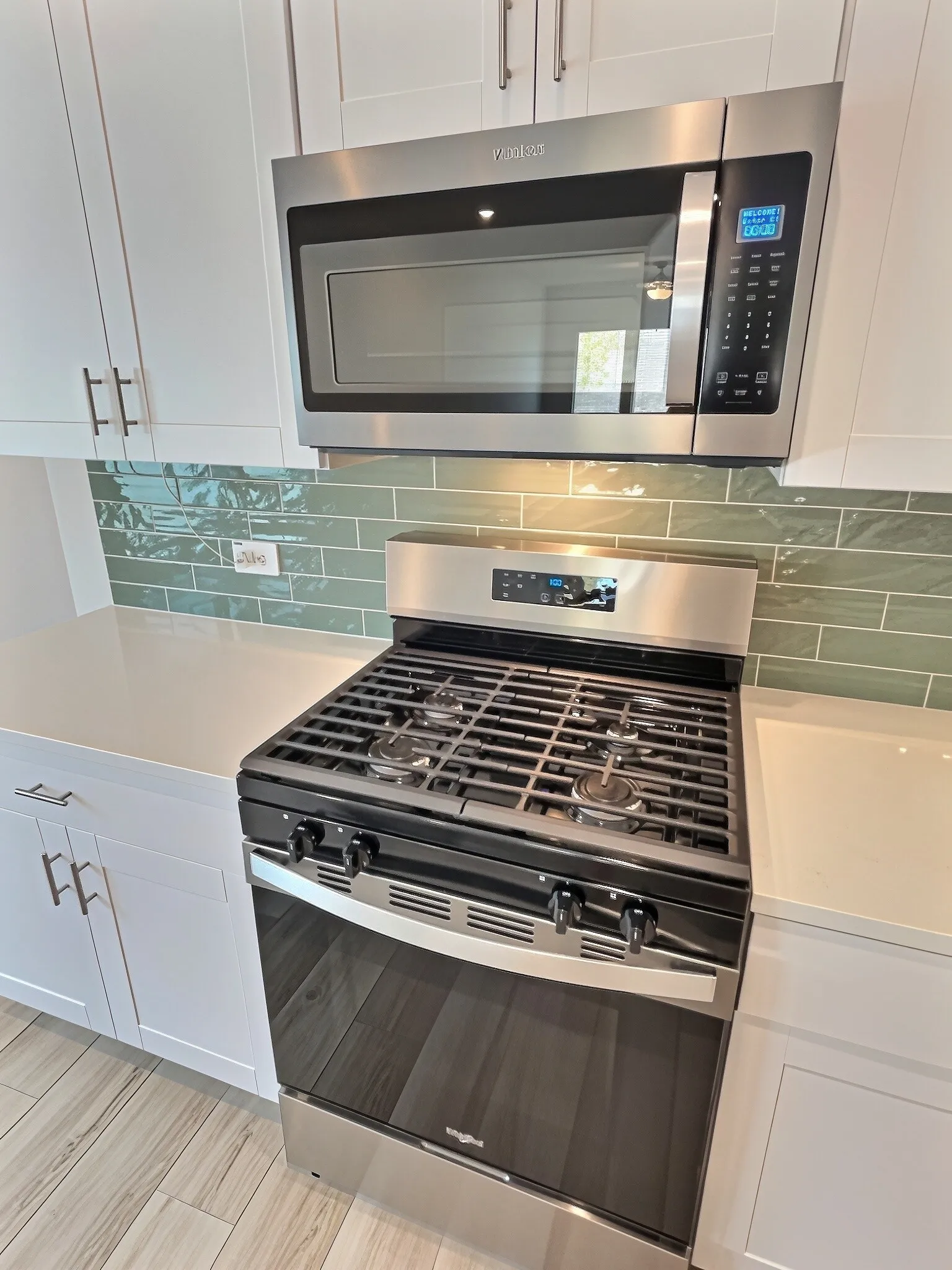 Stainless steel range with a built-in oven, coordinating microwave, white cabinetry with silver-tone hardware, white countertops, and a green subway tile backsplash