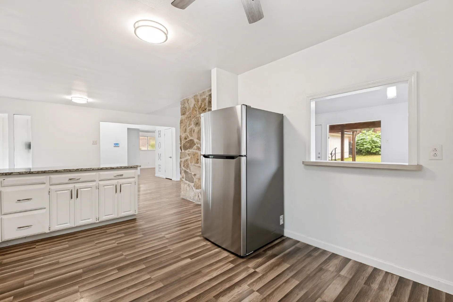 Spacious kitchen featuring white cabinetry with brushed nickel hardware, stainless steel refrigerator, wood-finish flooring, recessed and ceiling-mounted lighting, and a large window providing natural light