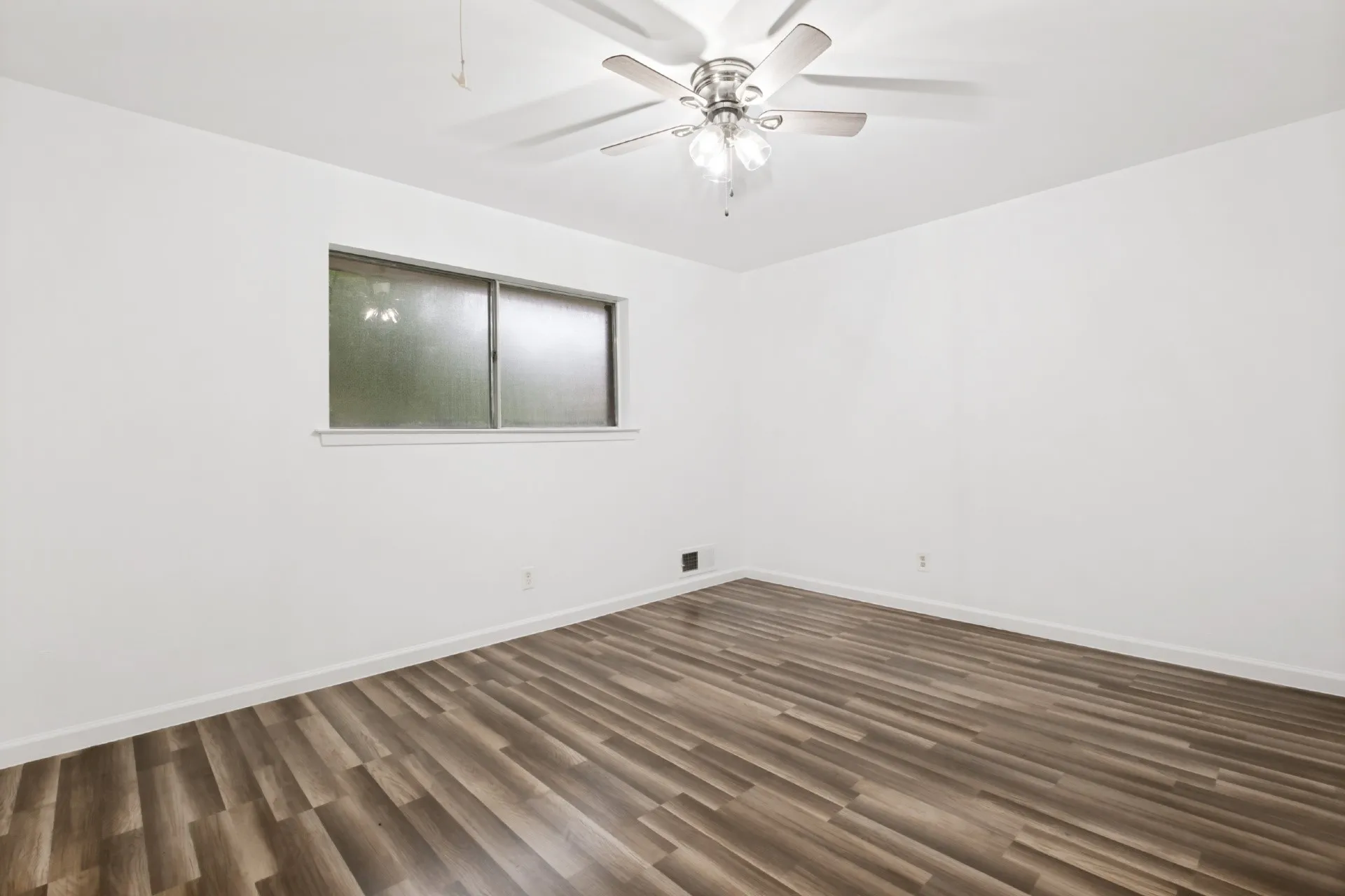 Spacious bedroom showcasing wood-finish flooring with linear plank detailing, white walls, ceiling fan with integrated lighting, and a horizontal window with dual-pane glass providing natural light