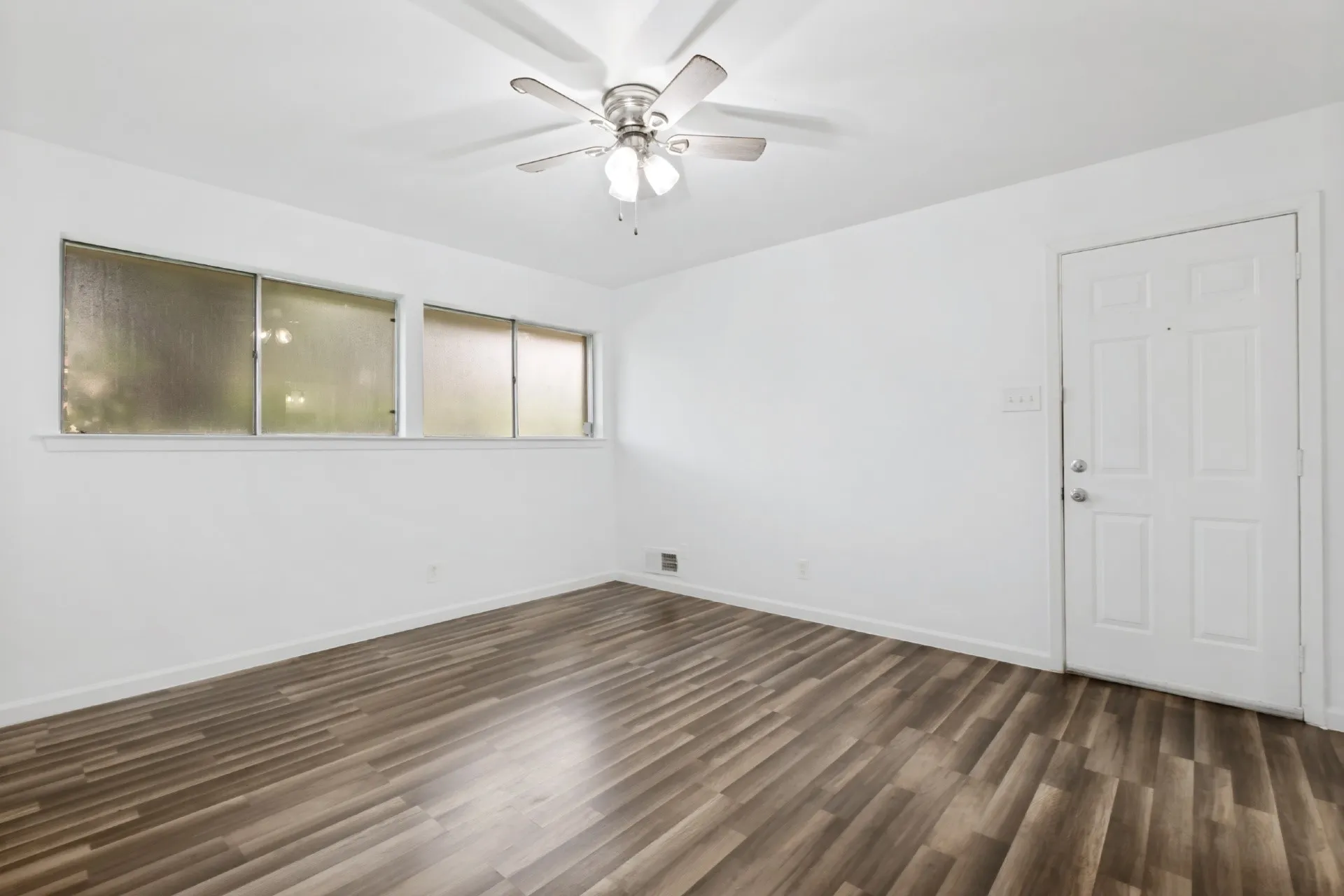 Wood-finish flooring, white walls, multiple windows, white paneled door, and a ceiling fan