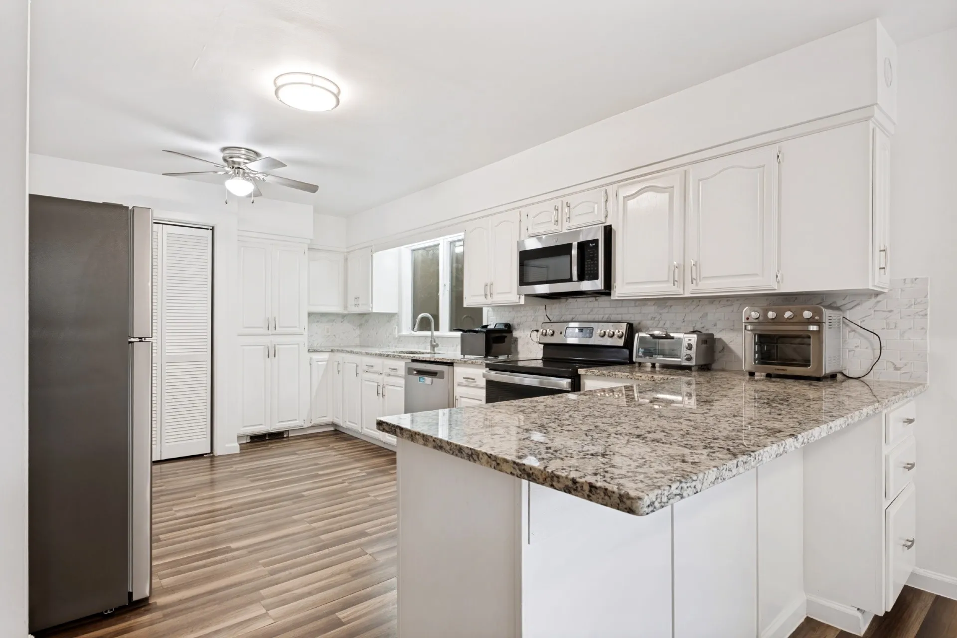 Modern kitchen featuring white cabinetry with ample storage, granite-style countertops with waterfall island edge, stainless steel appliances including gas range and microwave, and wood-finish flooring throughout the open layout