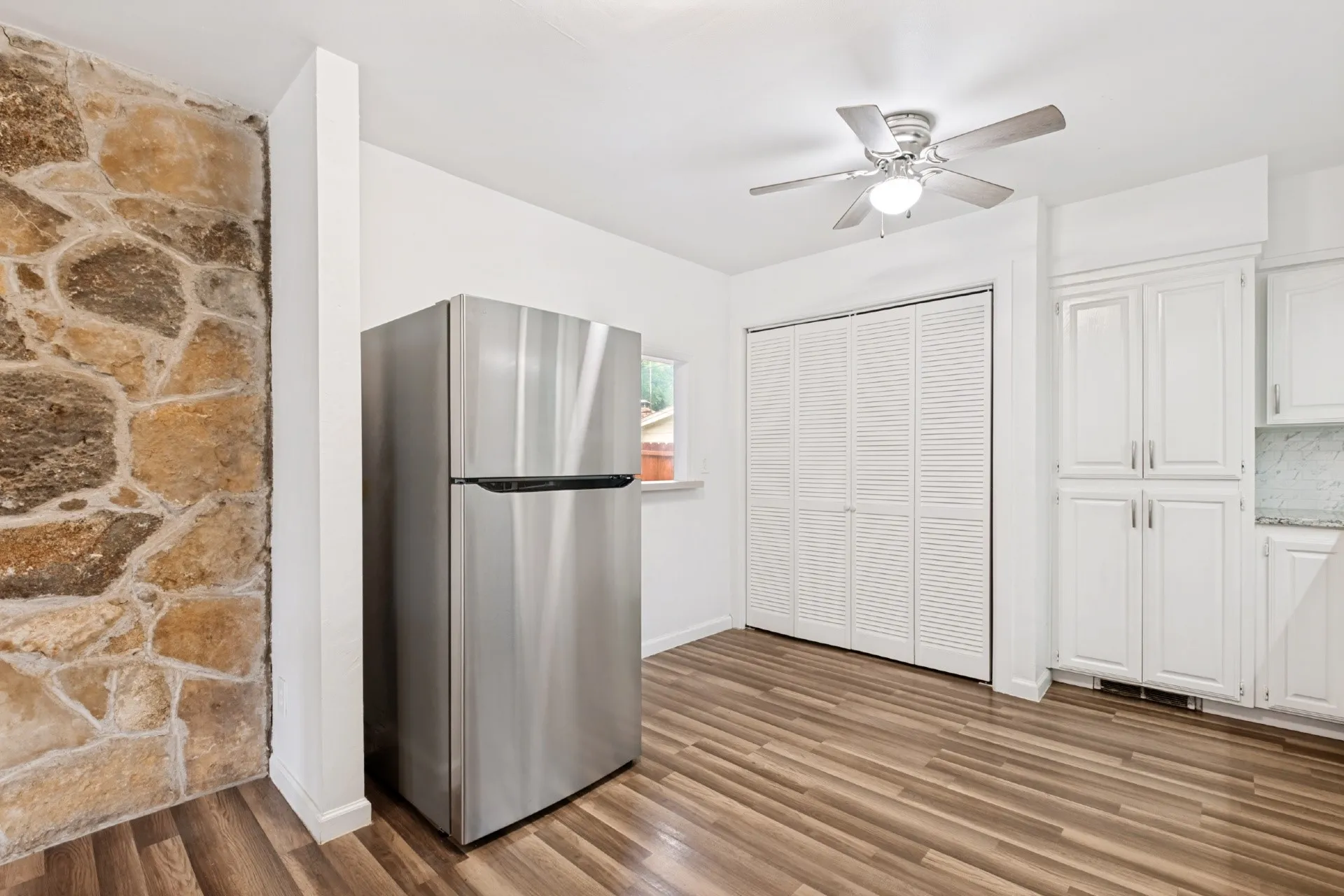 Stone accent wall, wood-finish flooring, stainless steel refrigerator, white shaker-style cabinetry, and ceiling fan with integrated lighting