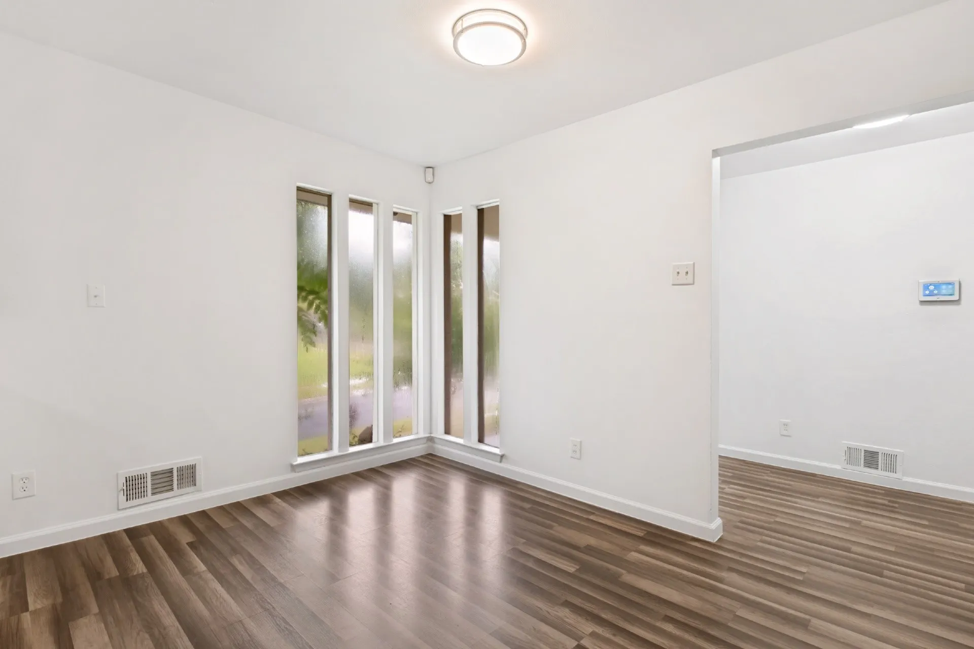 Bright corner room featuring expansive multi-pane windows with natural light, wood-finish flooring throughout, and clean white walls with minimal electrical outlets and light switches visible