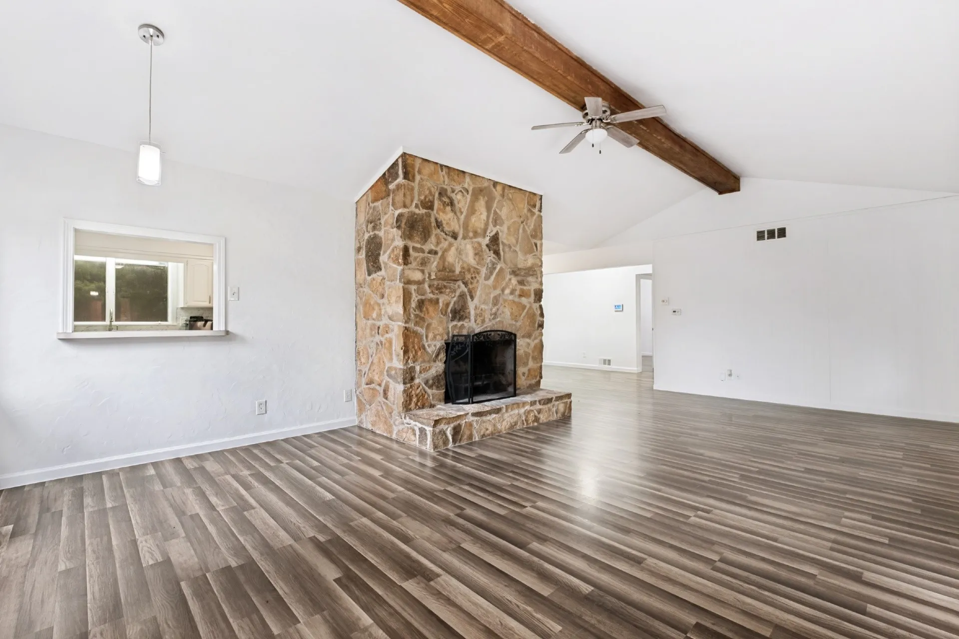Vaulted ceiling with exposed wood beam, wood-finish flooring, stone-clad fireplace with black grate, and a pass-through window