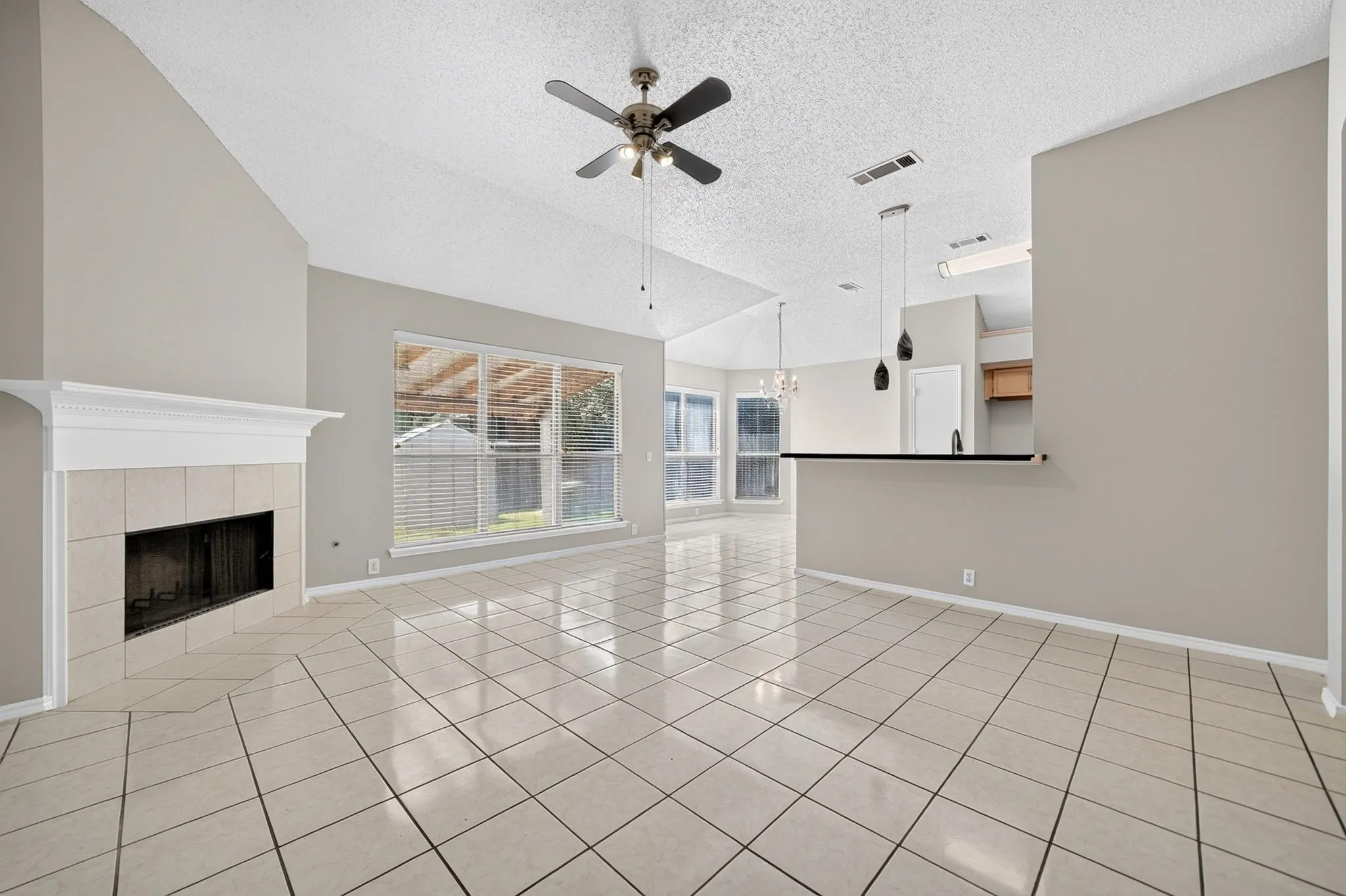 Vaulted ceiling with a ceiling fan, tiled flooring, fireplace with a mantel, large window with blinds, and a built-in breakfast bar with pendant lighting