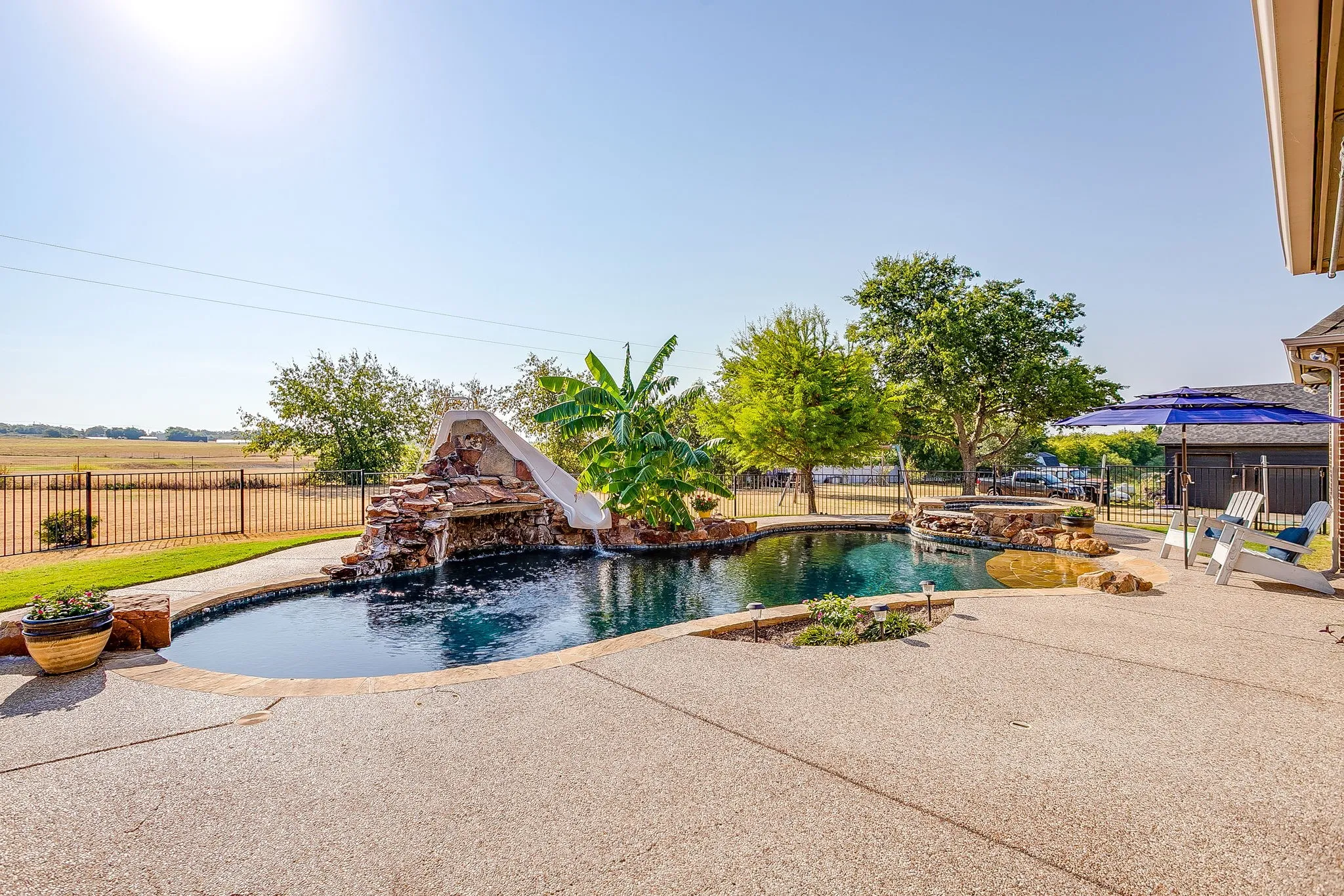 Curved swimming pool with a rock waterfall and slide, surrounded by an expansive textured concrete patio