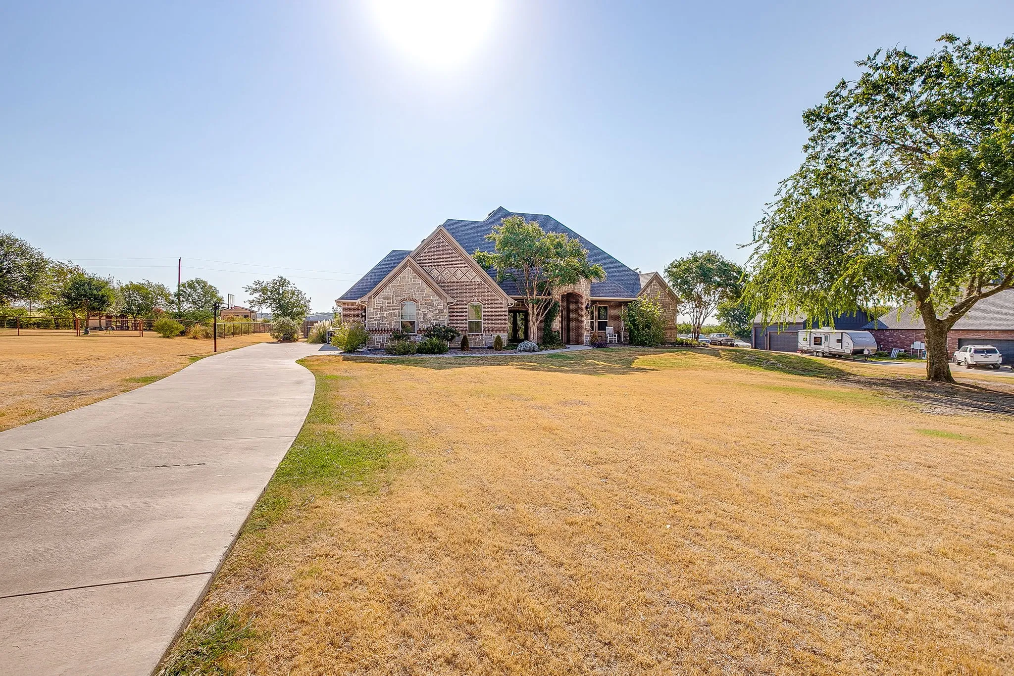 Brick and stone facade, expansive front lawn, mature trees, concrete drive, and a covered entry