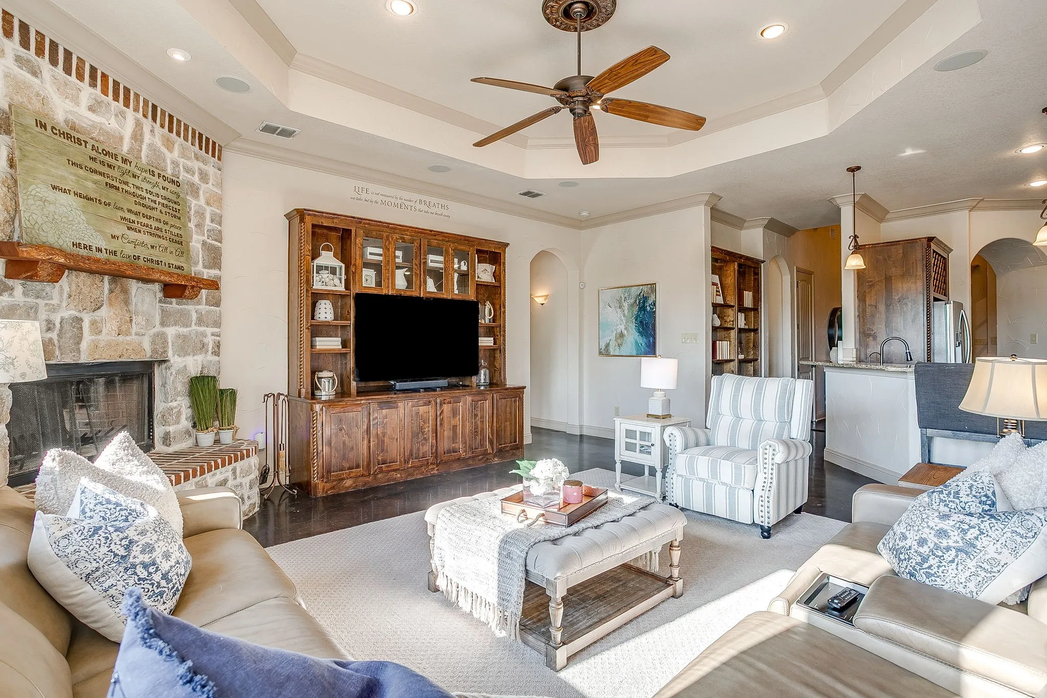 Stone fireplace with a wood-beam mantel, built-in wooden entertainment center, tray ceiling with a ceiling fan, dark wood-finish flooring, and recessed lighting