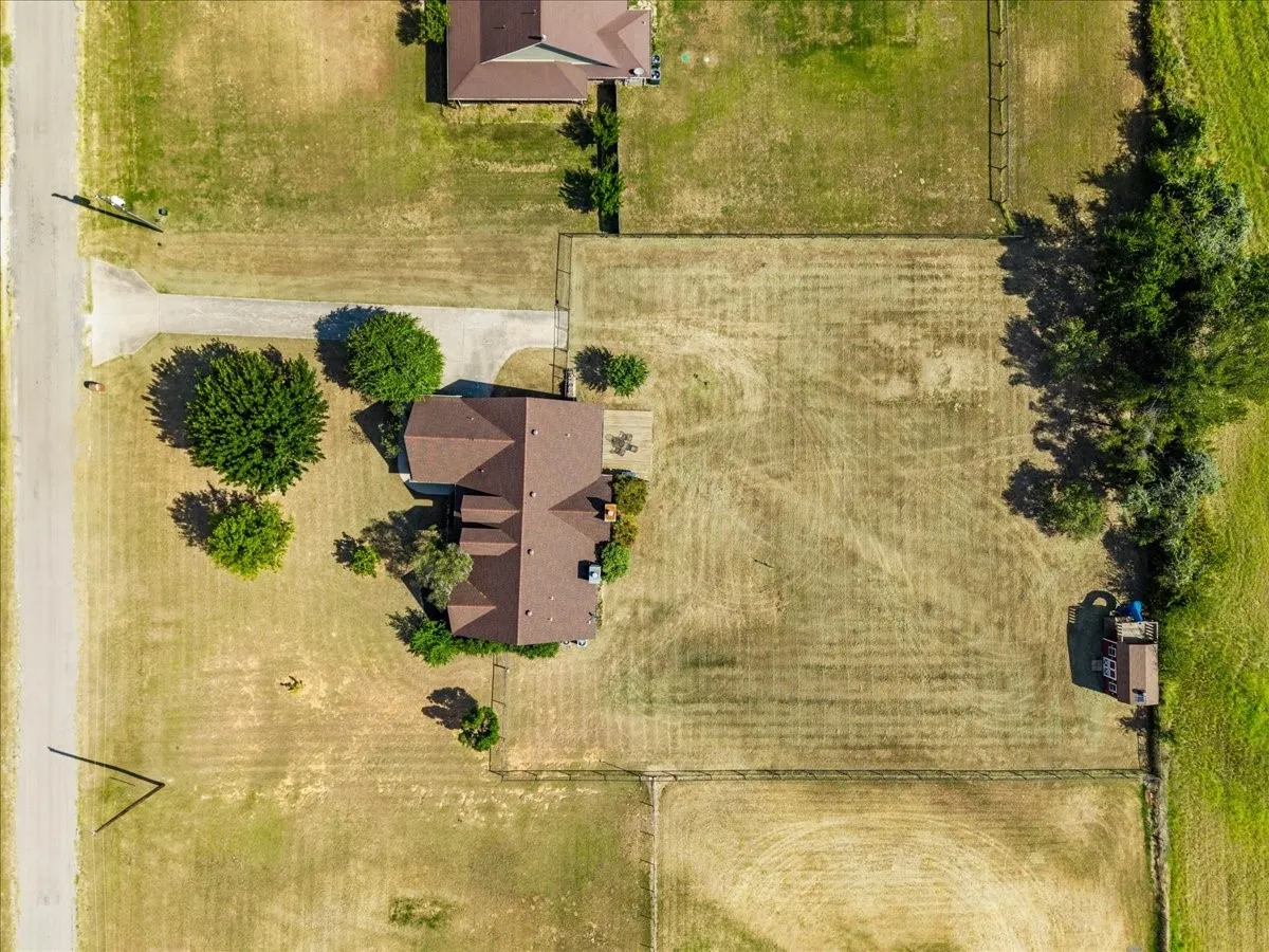 Aerial property view featuring a brown shingle roof, mature trees, an asphalt driveway, and fenced perimeter