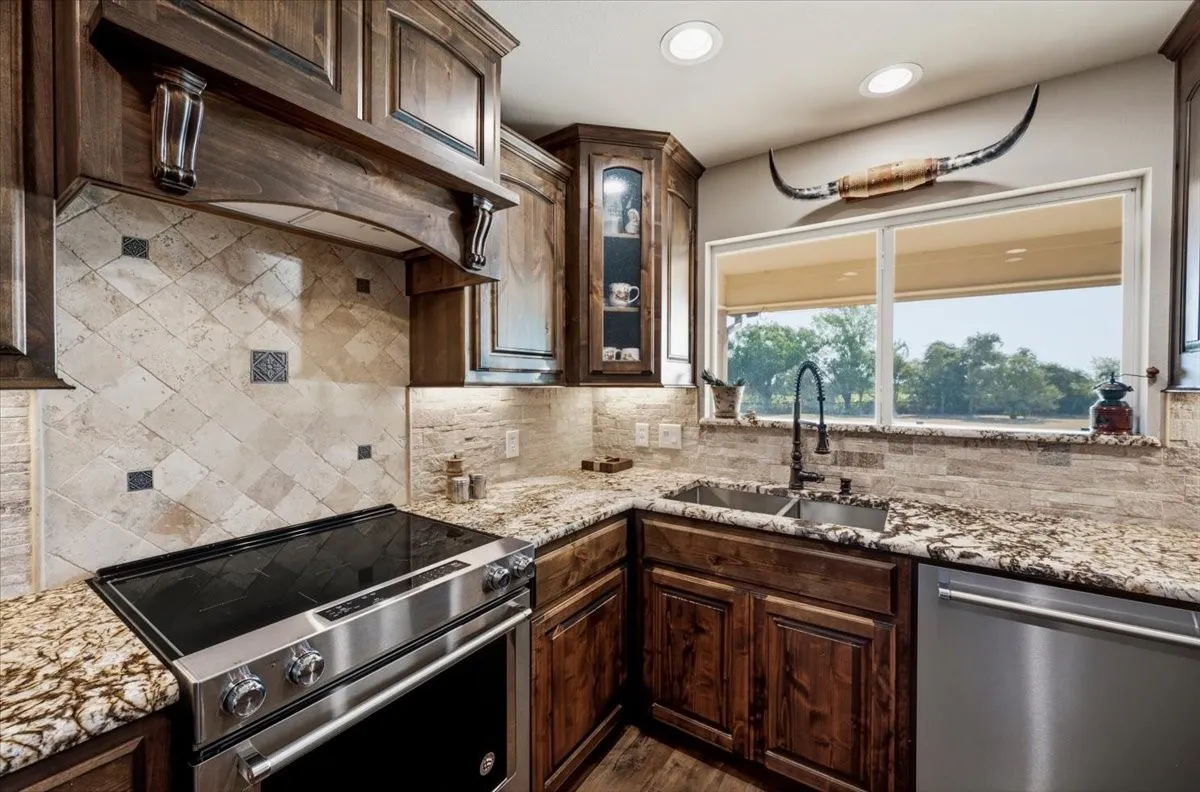 Stainless steel range, wood cabinetry, stone countertops, stone tile backsplash, and a double basin sink with a gooseneck faucet
