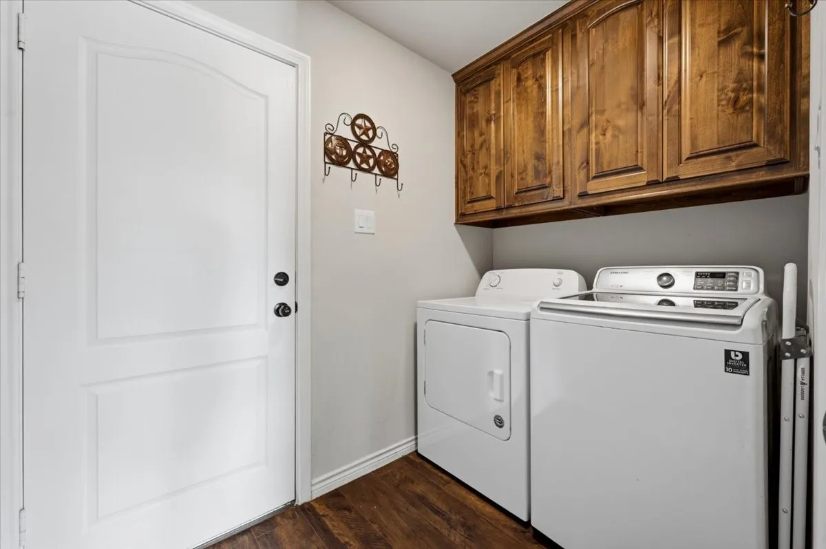 Dedicated laundry room featuring side-by-side washer and dryer with digital controls, rich wood-finish cabinetry with ample storage above, warm wood-finish flooring, and convenient utility sink for added functionality