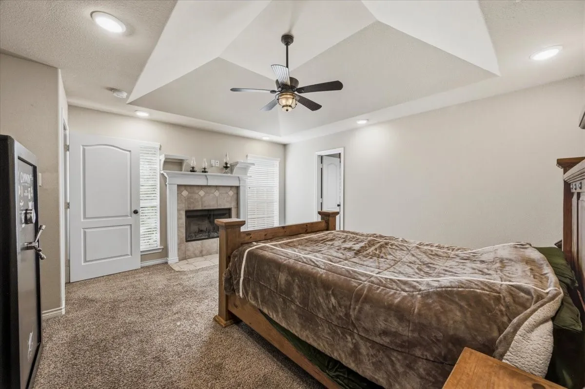 Spacious primary bedroom suite showcasing a coffered ceiling with recessed lighting, ceiling fan with integrated fixture, and a gas fireplace with white surround and stone accents