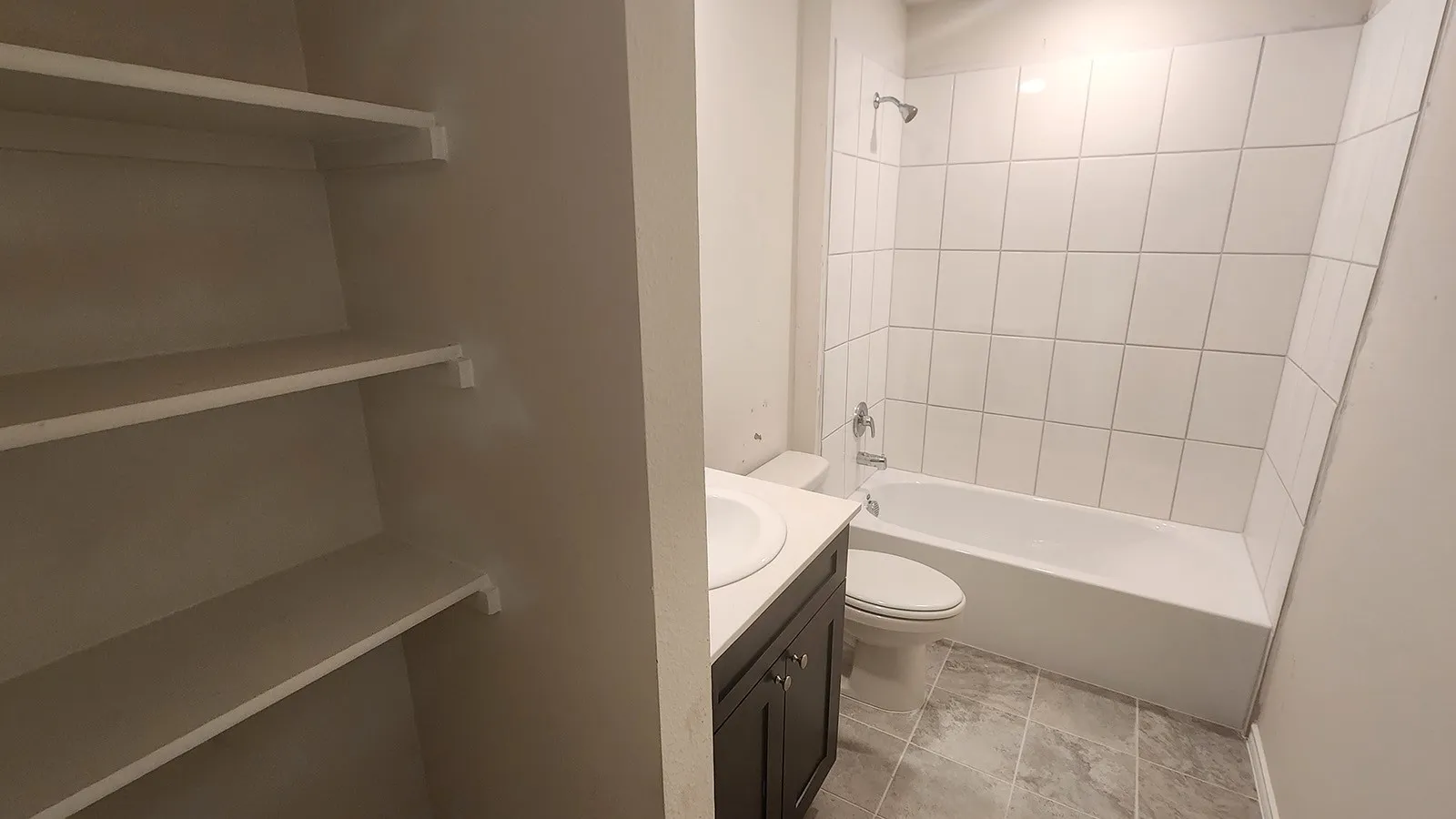 Built-in shelving, dark wood-finish vanity with countertop, undermount sink, white tile shower surround, and gray-toned floor tiling