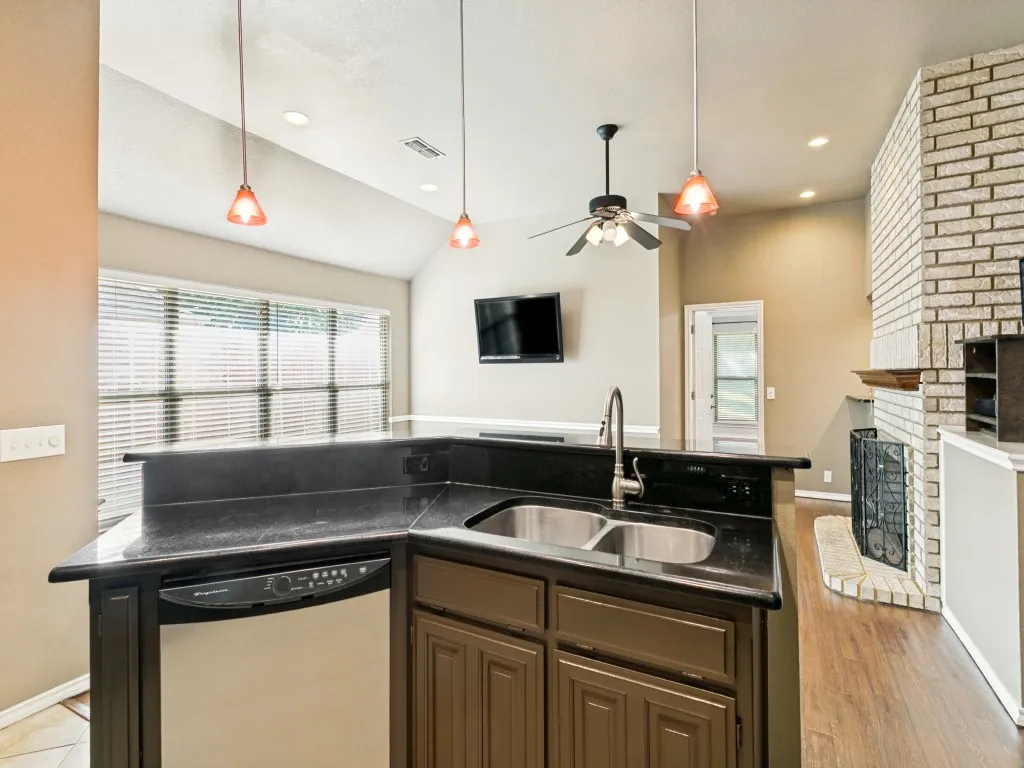 Kitchen island with a dark countertop and double basin sink, integrated dishwasher, adjacent brick-face fireplace, and wood-finish flooring
