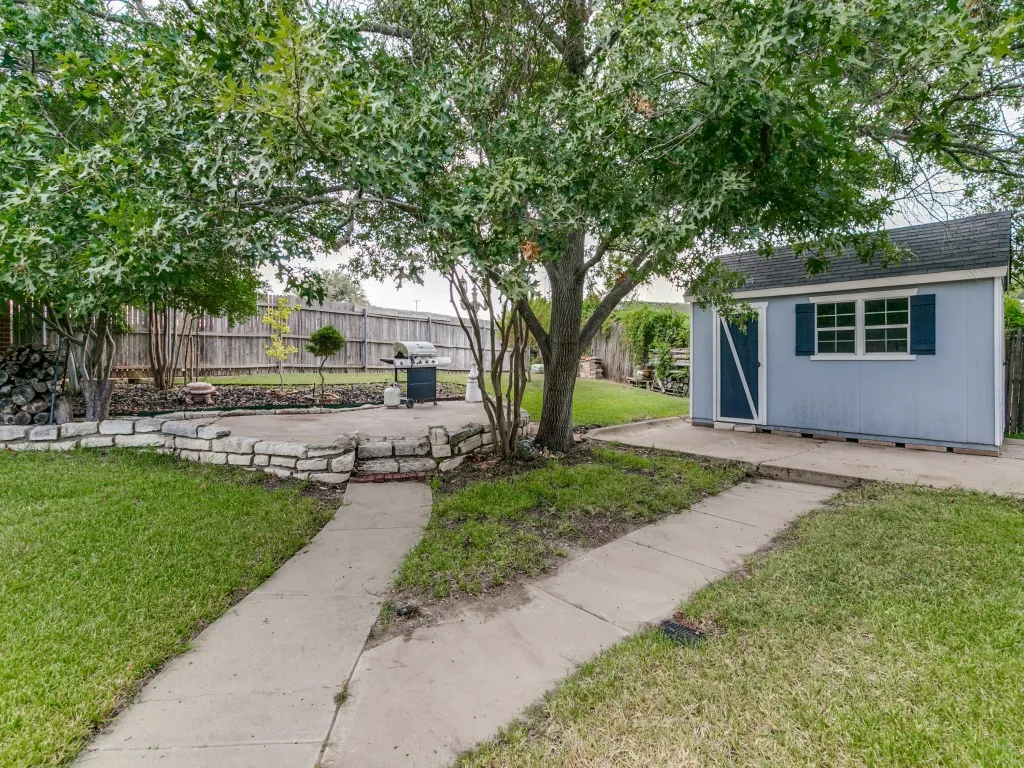 Expansive backyard featuring a stone-lined patio, mature trees, a storage shed with shutters, and a privacy fence