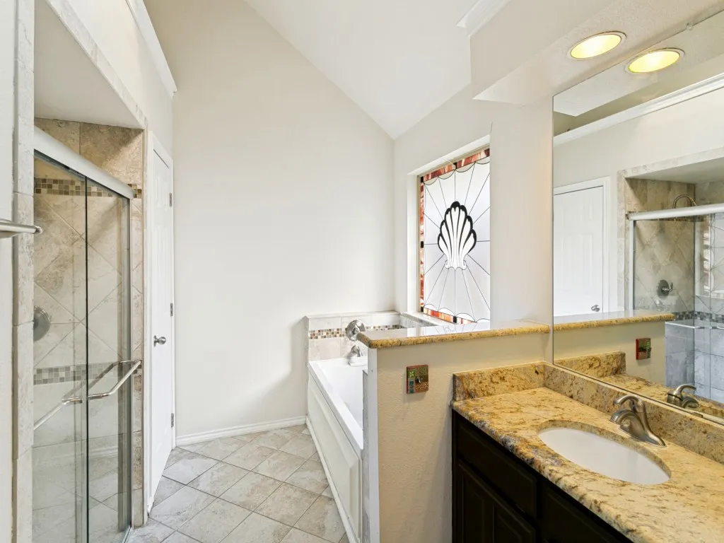 Angled vaulted ceiling, freestanding bathtub, stained glass window, tiled shower with glass enclosure, and a vanity with an oval sink and stone countertop