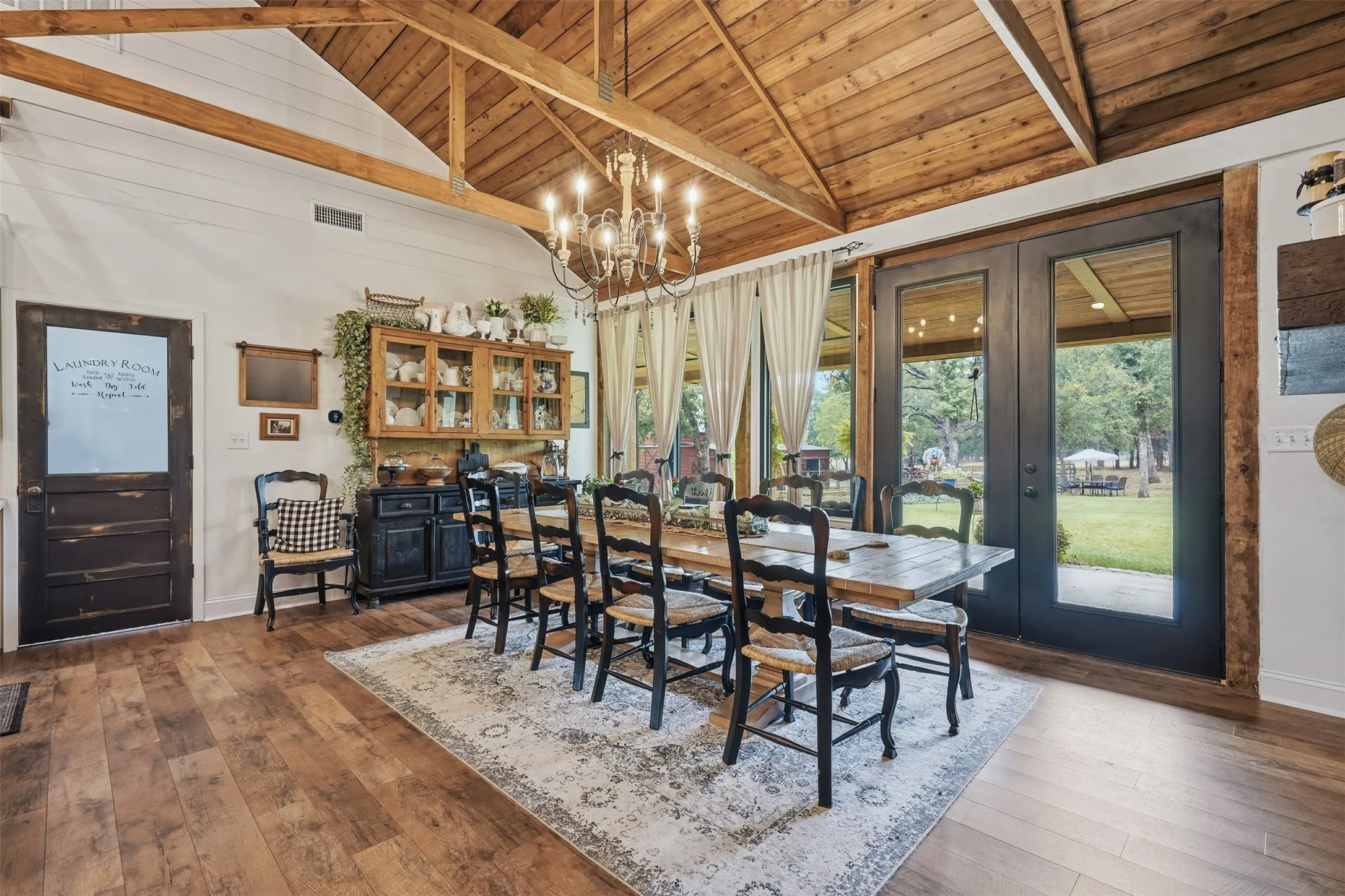 vaulted wood ceiling with multiple chandeliers and glass looking out to tree shaded lot and covered porches