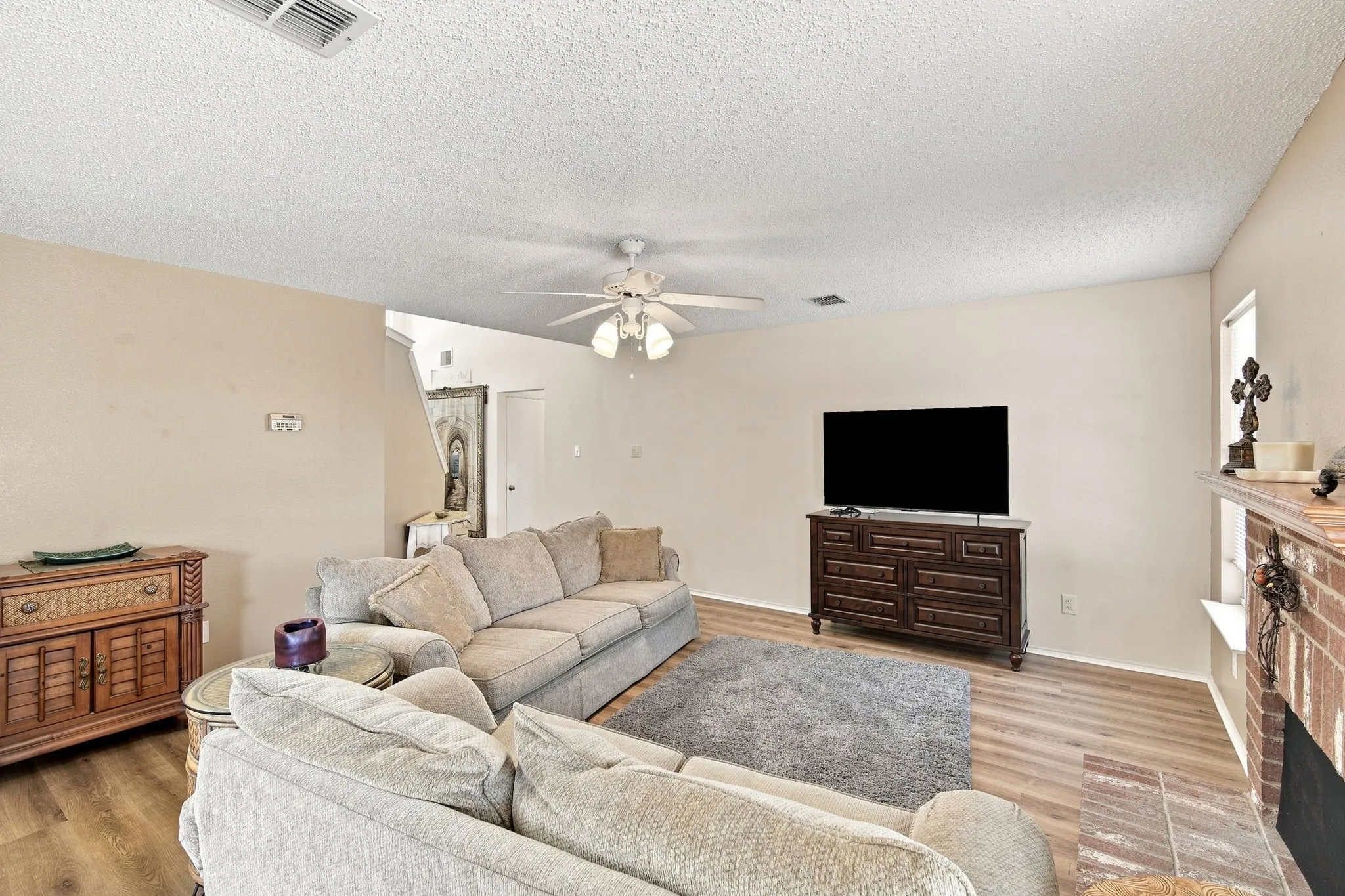 Wood-finish flooring, ceiling fan with integrated lighting, and a brick-faced fireplace with a mantel