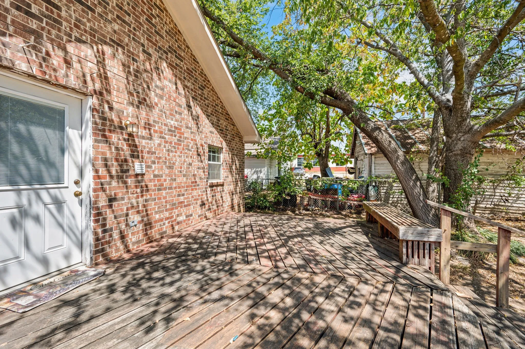 Brick exterior with a white door, large shade tree, and a wooden deck with an integrated bench