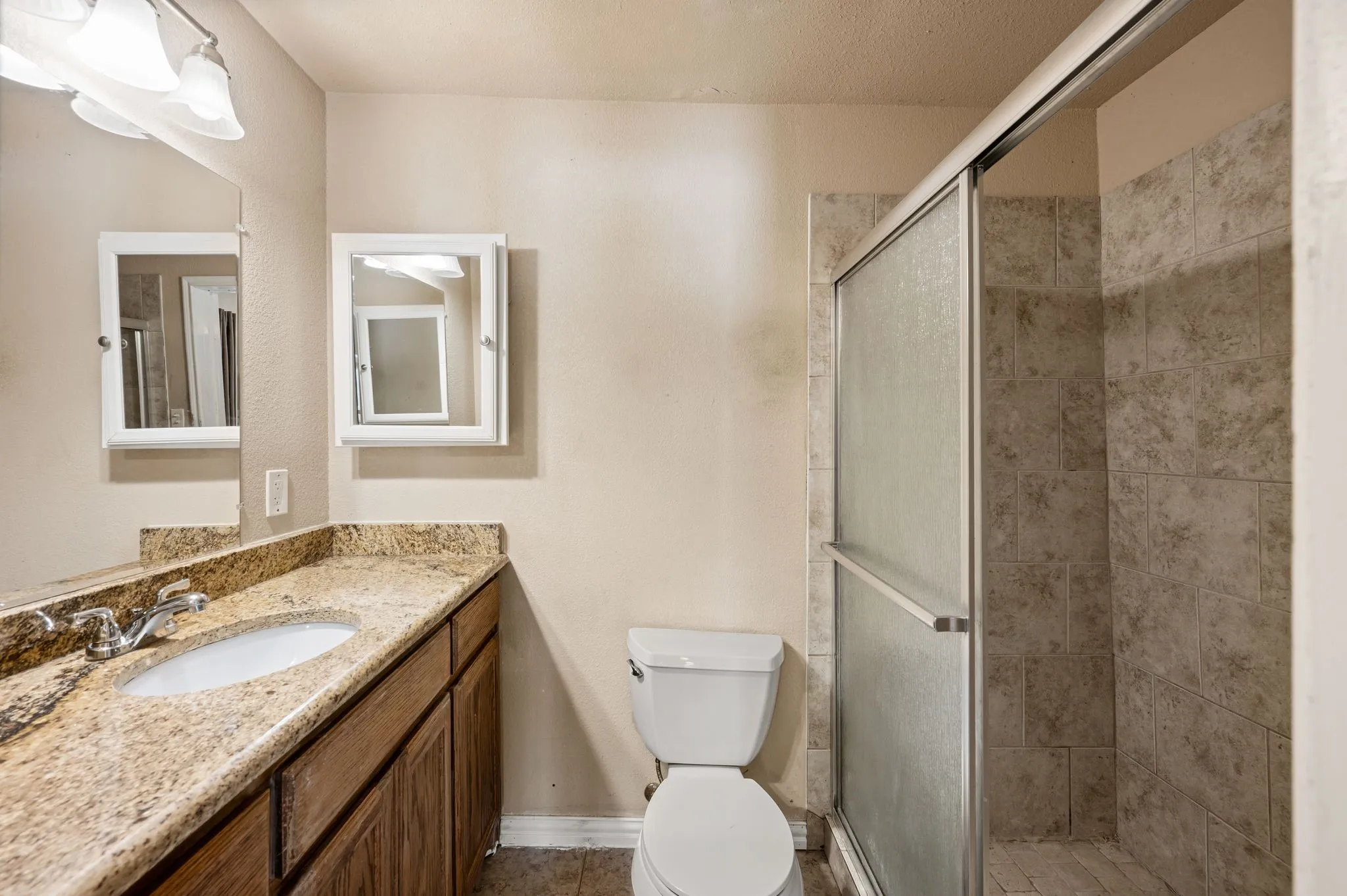 Vanity with an integrated sink and stone countertop, wood-finish cabinetry, two mirrored medicine cabinets, and a tiled shower with a sliding glass door