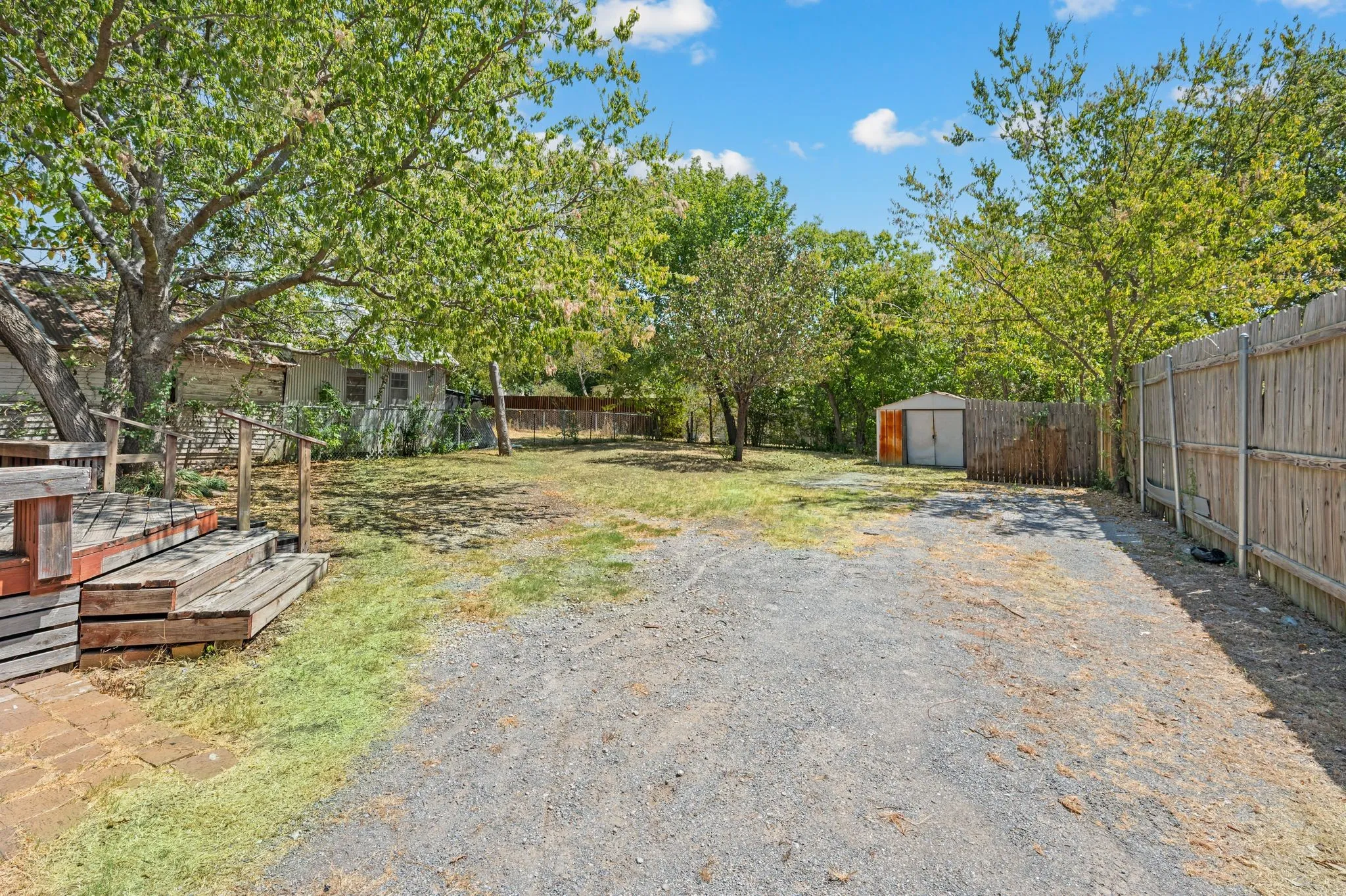 Spacious exterior featuring a gravel driveway, wood deck, mature trees, and a metal shed