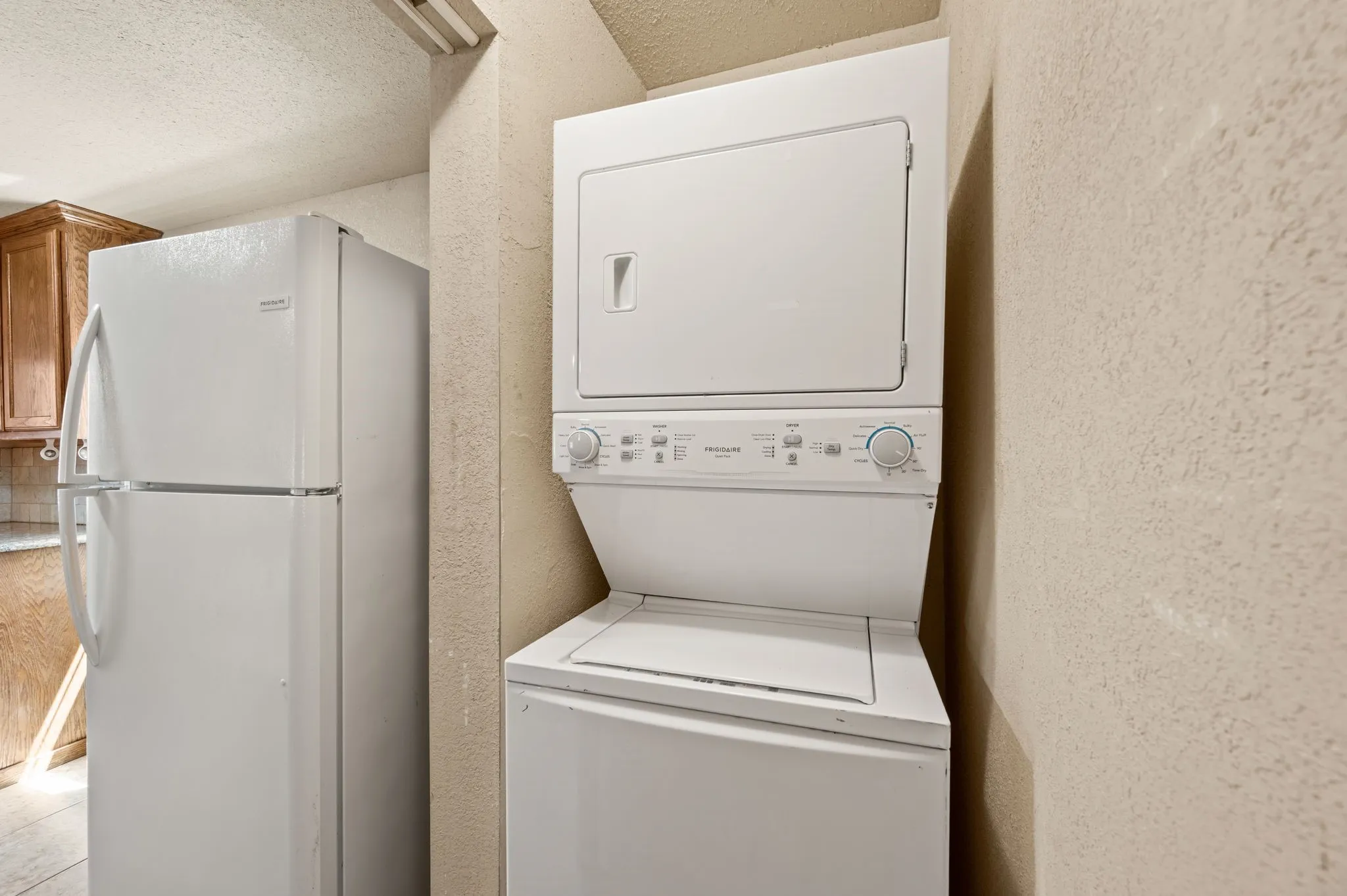 White stackable washer and dryer, white refrigerator, and wood-finish cabinetry with light-toned countertops