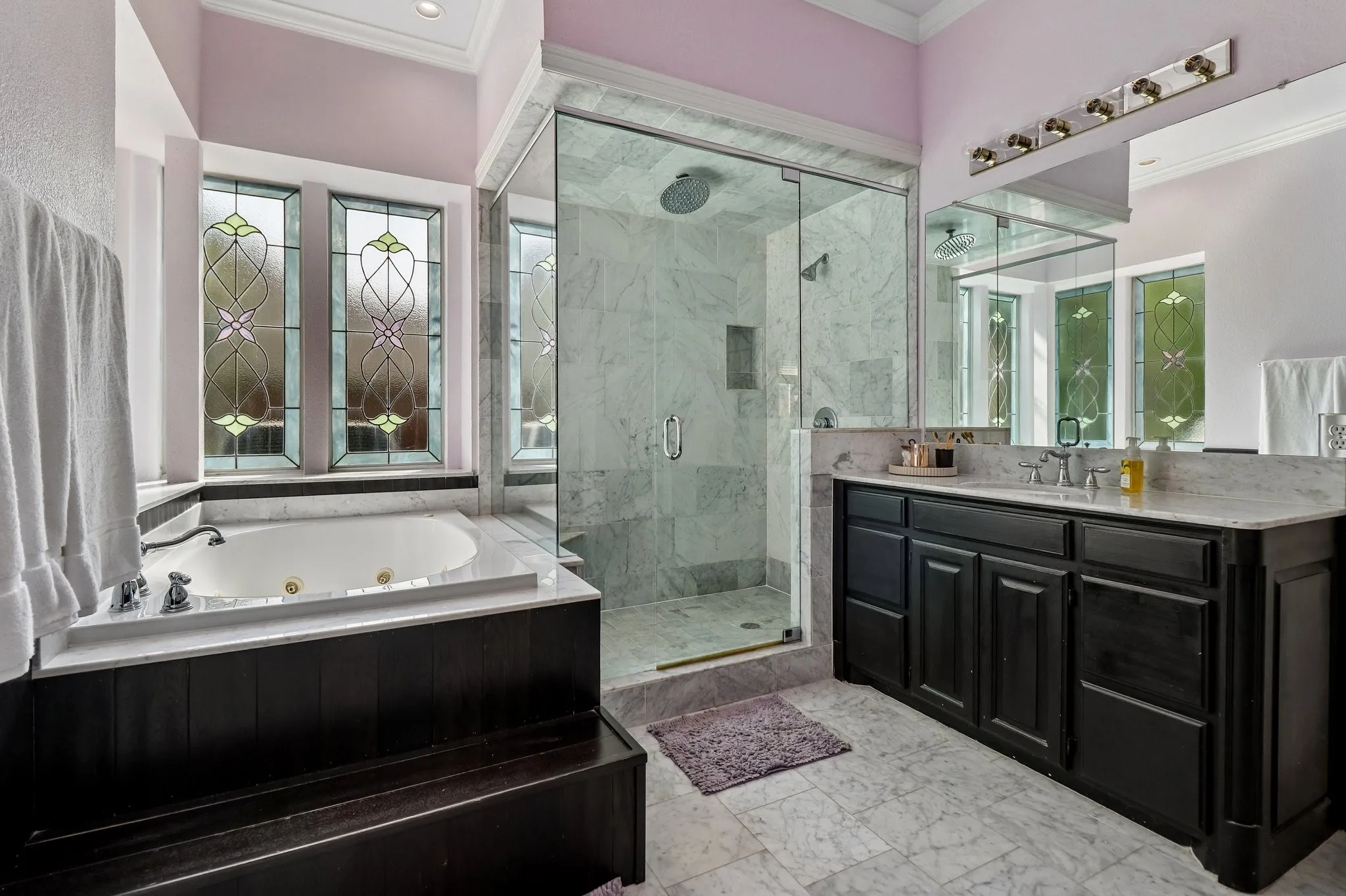 Glass-enclosed shower with a rainfall showerhead, jetted soaking tub, dark wood-finish vanity with a light-toned countertop, and patterned leaded glass windows
