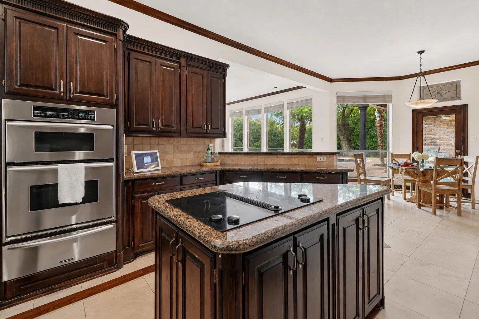 Built-in double ovens, dark wood cabinetry, a center island with cooktop, granite countertops, and tile flooring