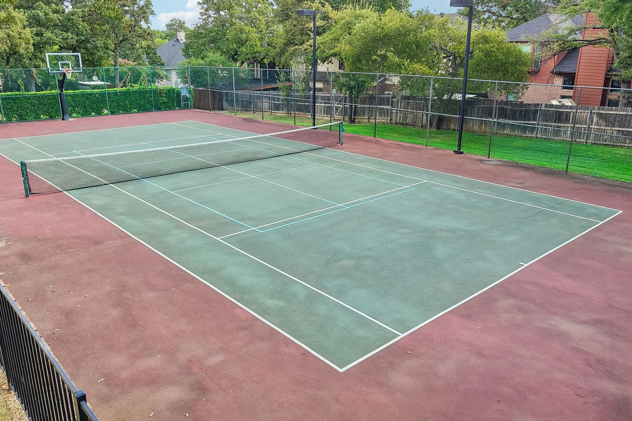 Full-size tennis court featuring a green playing surface with white line markings, surrounded by a red-toned court perimeter