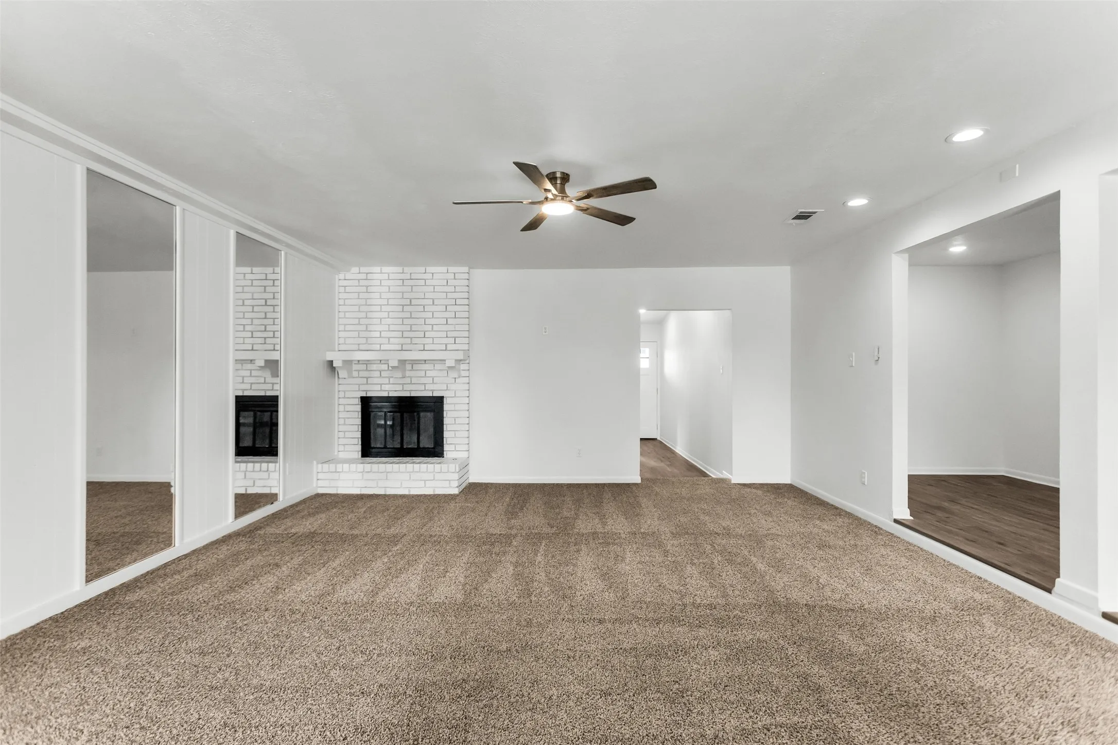 Spacious room with plush carpet flooring, a white brick fireplace, mirrored wall paneling, and a ceiling fan with integrated lighting