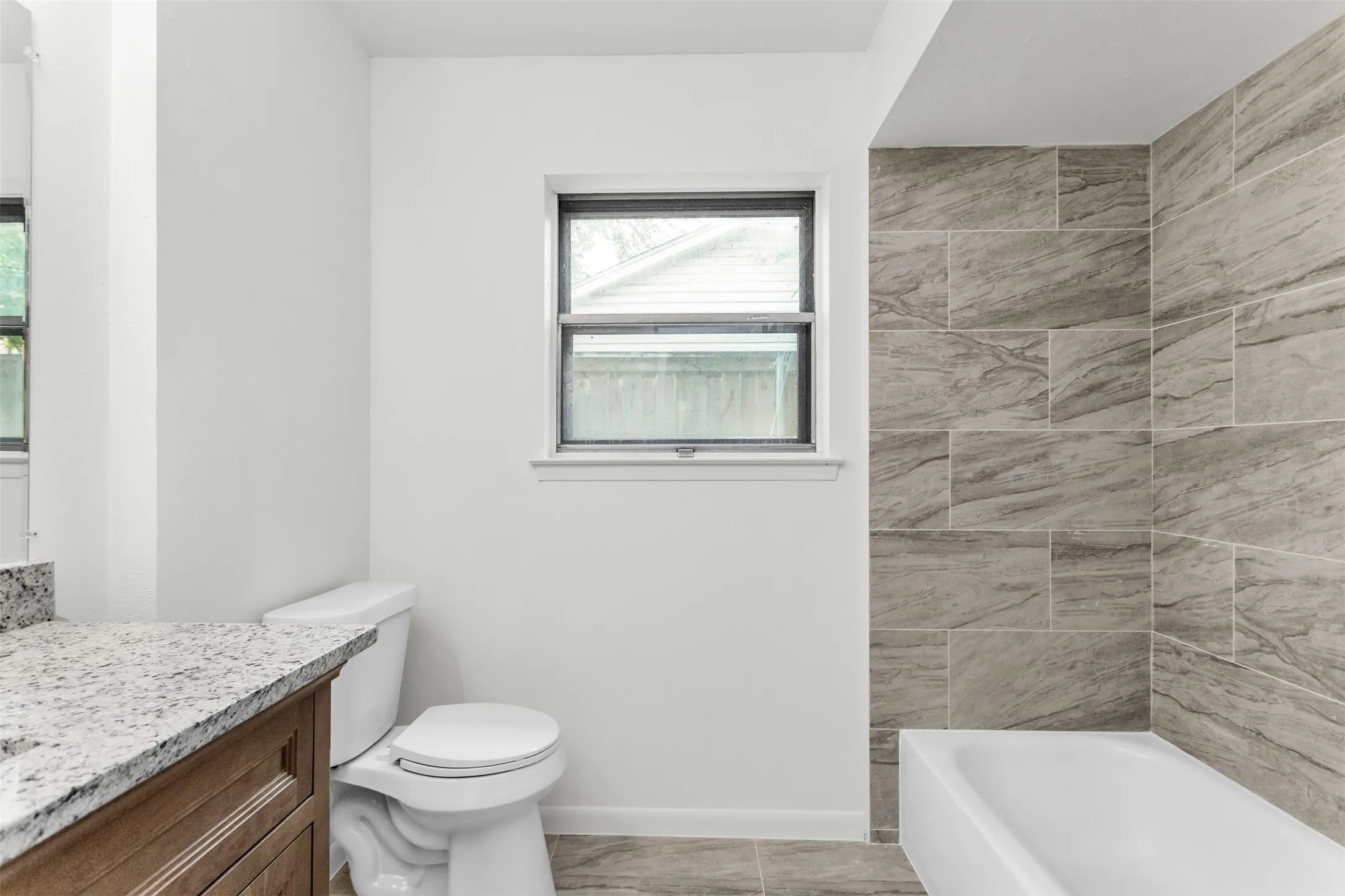 White bathtub with large format stone tile surround, a white toilet, and a window with a dark frame