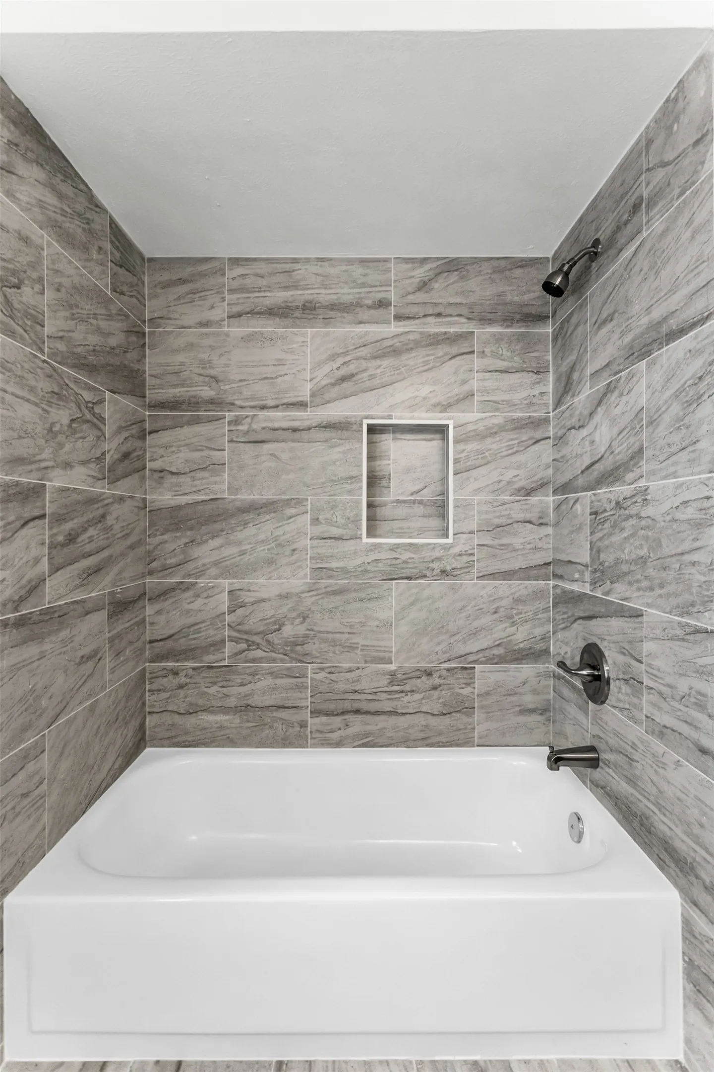 Bathtub with gray rectangular wall tiles, a built-in shower niche, and a dark bronze shower fixture