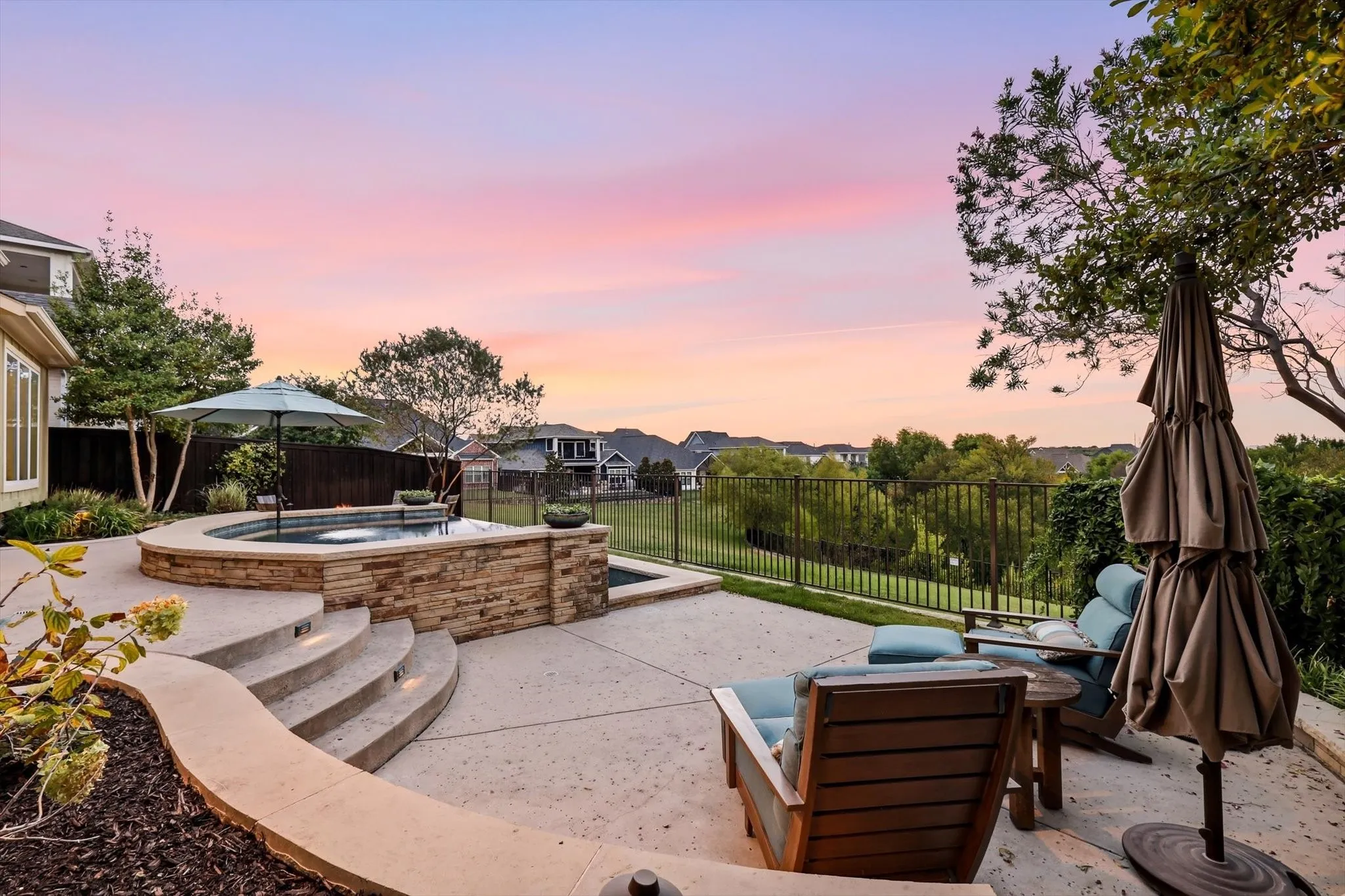 Outdoor patio with a built-in spa, stone-clad retaining wall, concrete steps with integrated lighting, and a black metal fence