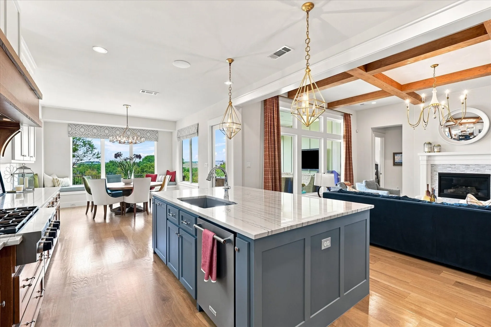 Open-concept interior featuring wood-finish flooring, a kitchen island with an undermount sink, a gas range with an oven, a built-in fireplace, and exposed wood beam ceiling details