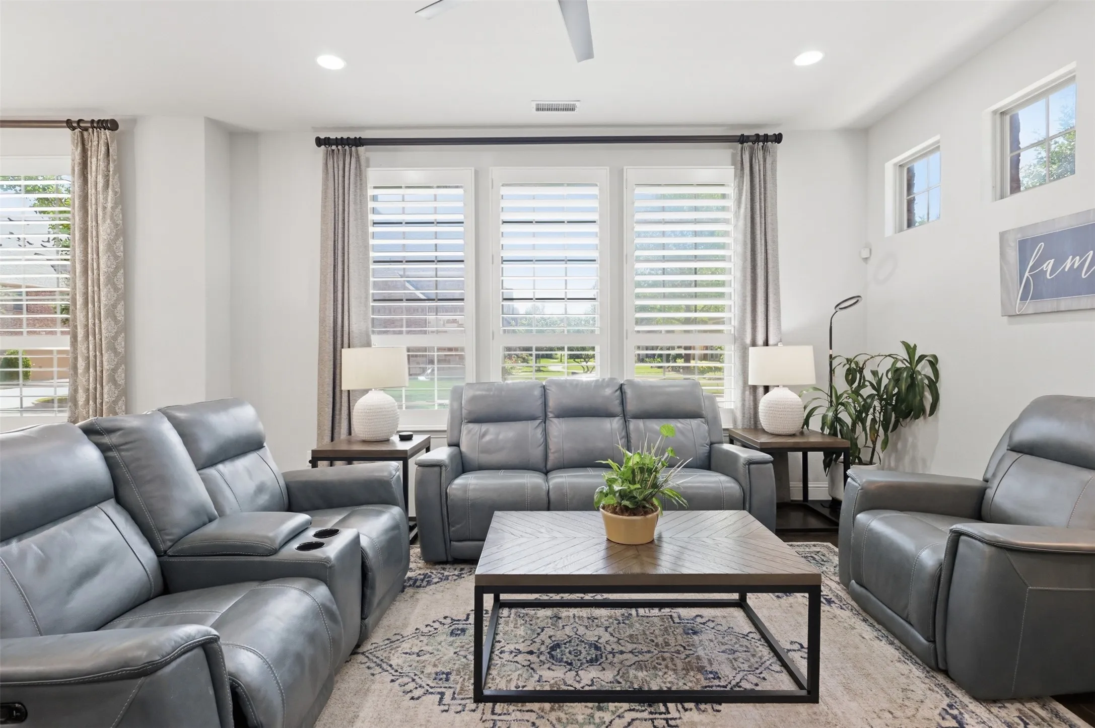 Sophisticated living room featuring expansive windows with plantation shutters and natural light, neutral color palette with recessed ceiling lighting, plush gray leather seating arrangements, and a contemporary coffee table anchoring the space