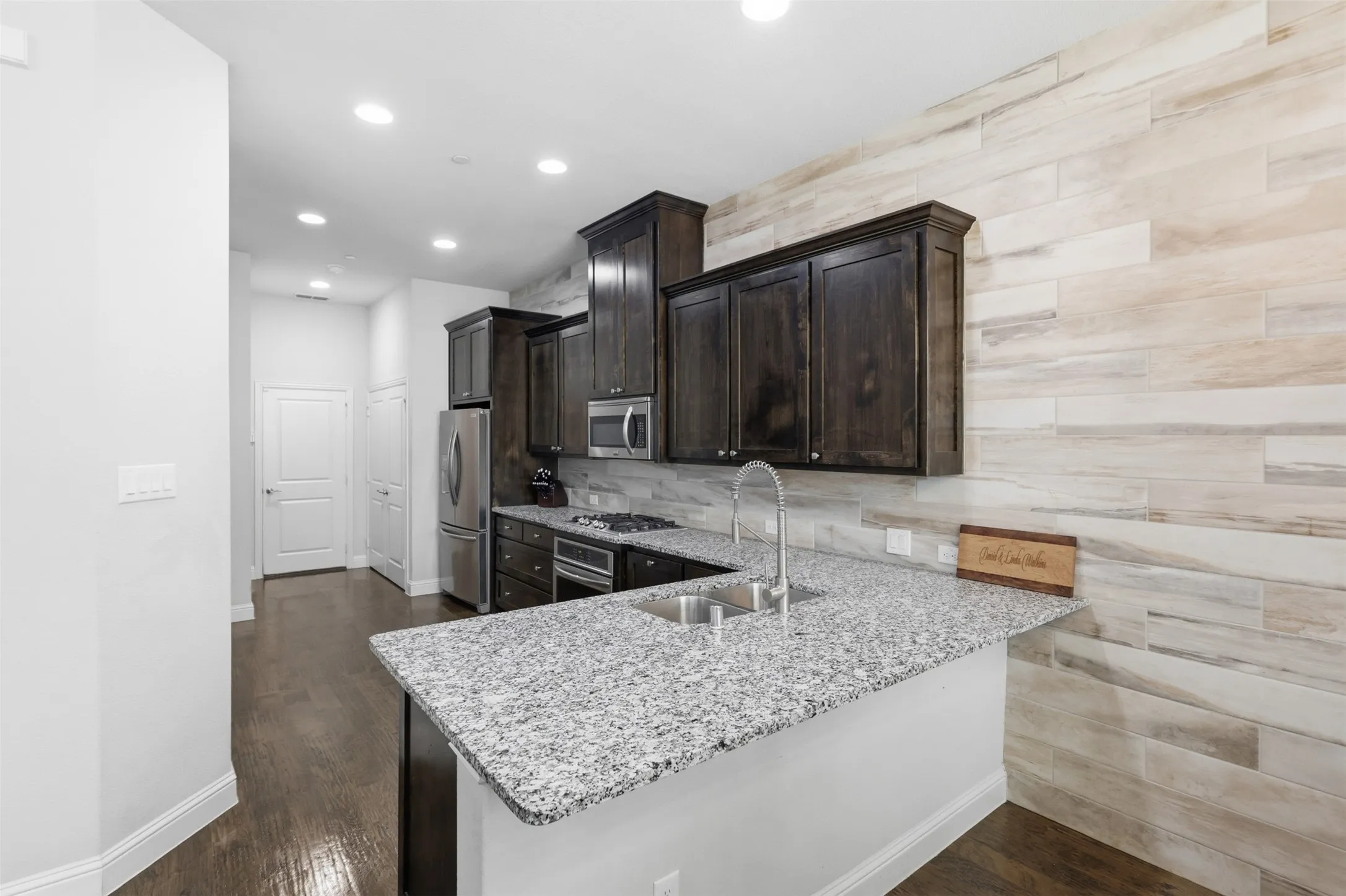 Kitchen featuring granite countertops, wood-finish flooring, dark wood cabinetry, stainless steel appliances, and a tiled accent wall