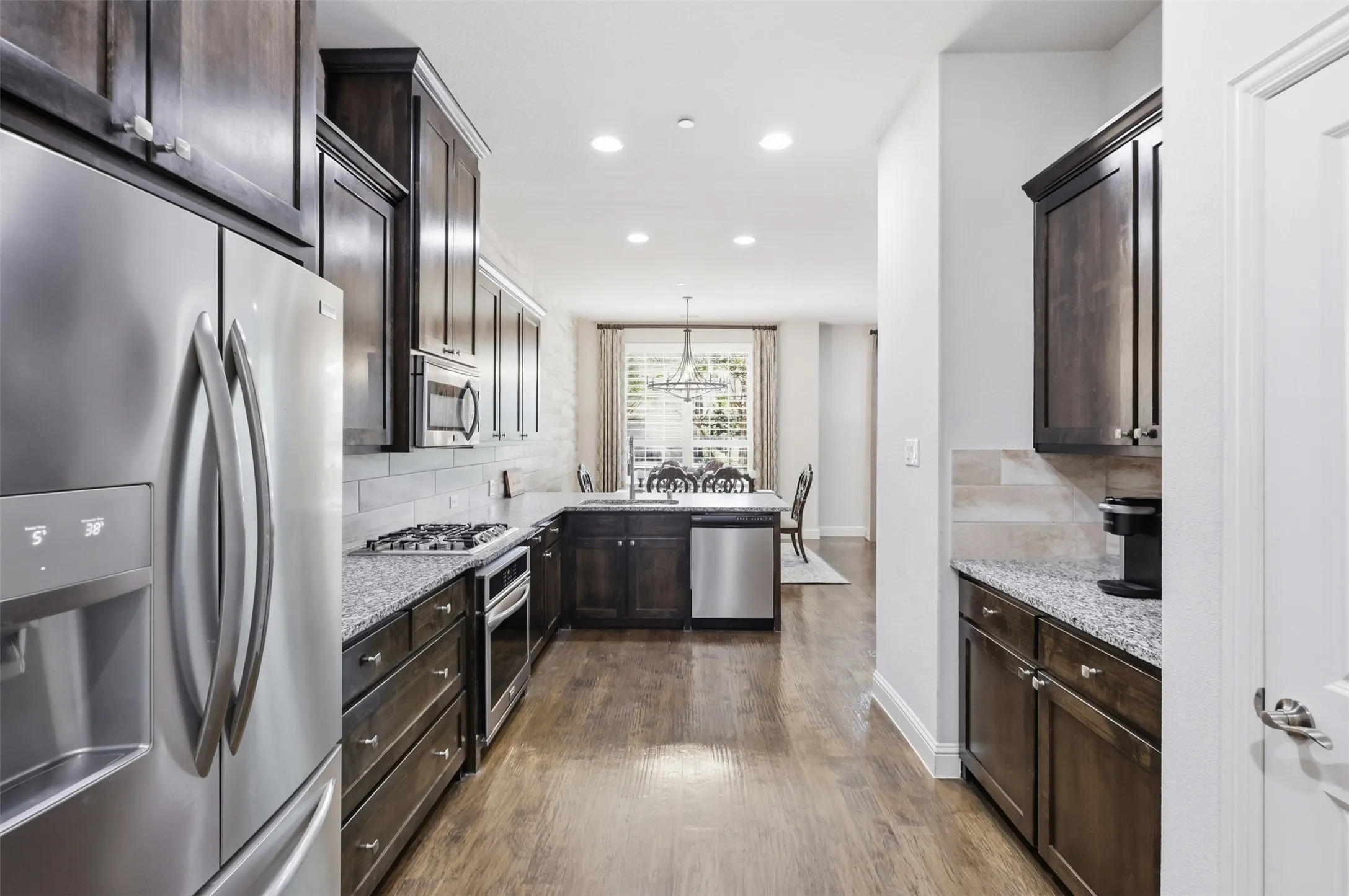 Stainless steel appliances, wood-finish flooring, dark wood cabinetry, light-colored backsplash, recessed lighting
