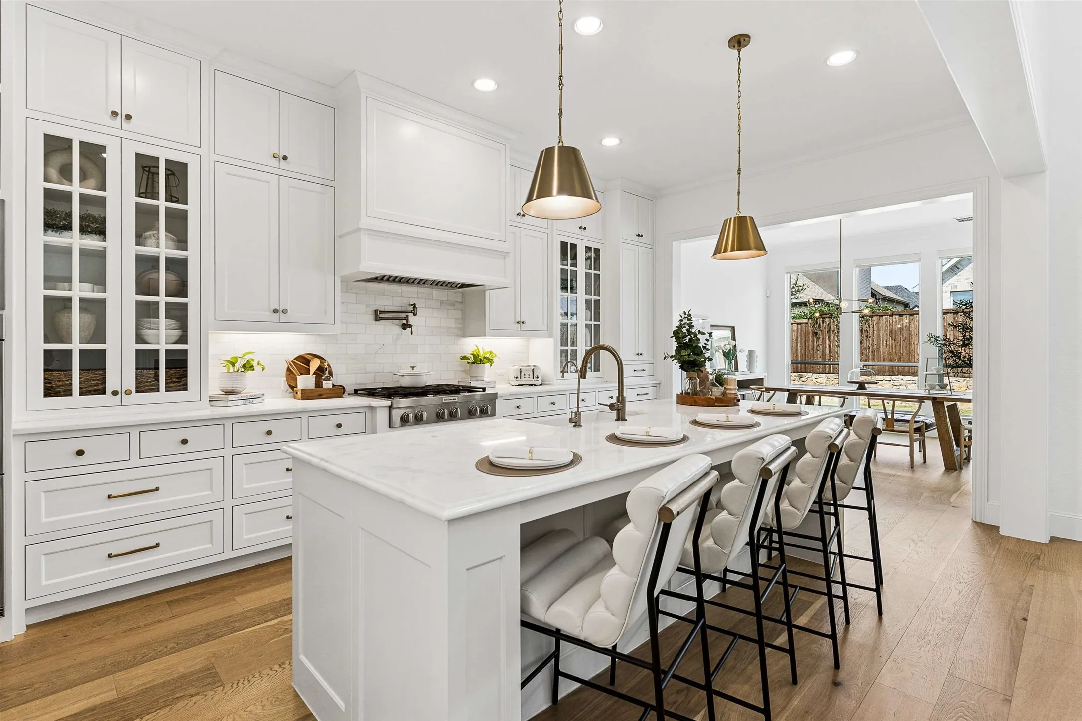 White shaker-style cabinetry, a substantial kitchen island with seating, pendant lighting, wood-finish flooring, and a large window offering natural light