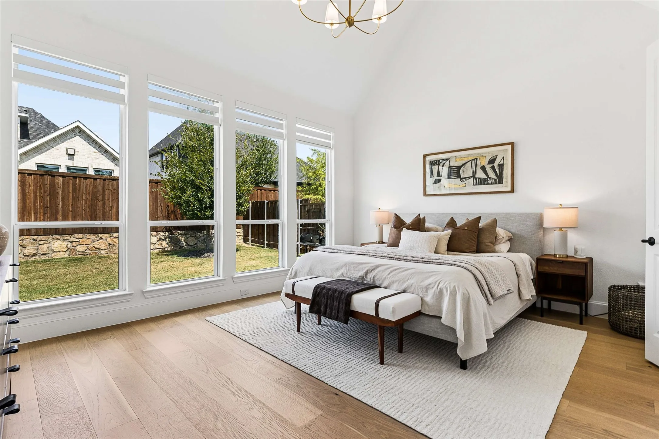 Vaulted ceiling with an elegant chandelier, expansive windows, wood-finish flooring, and a fenced yard with stone landscaping