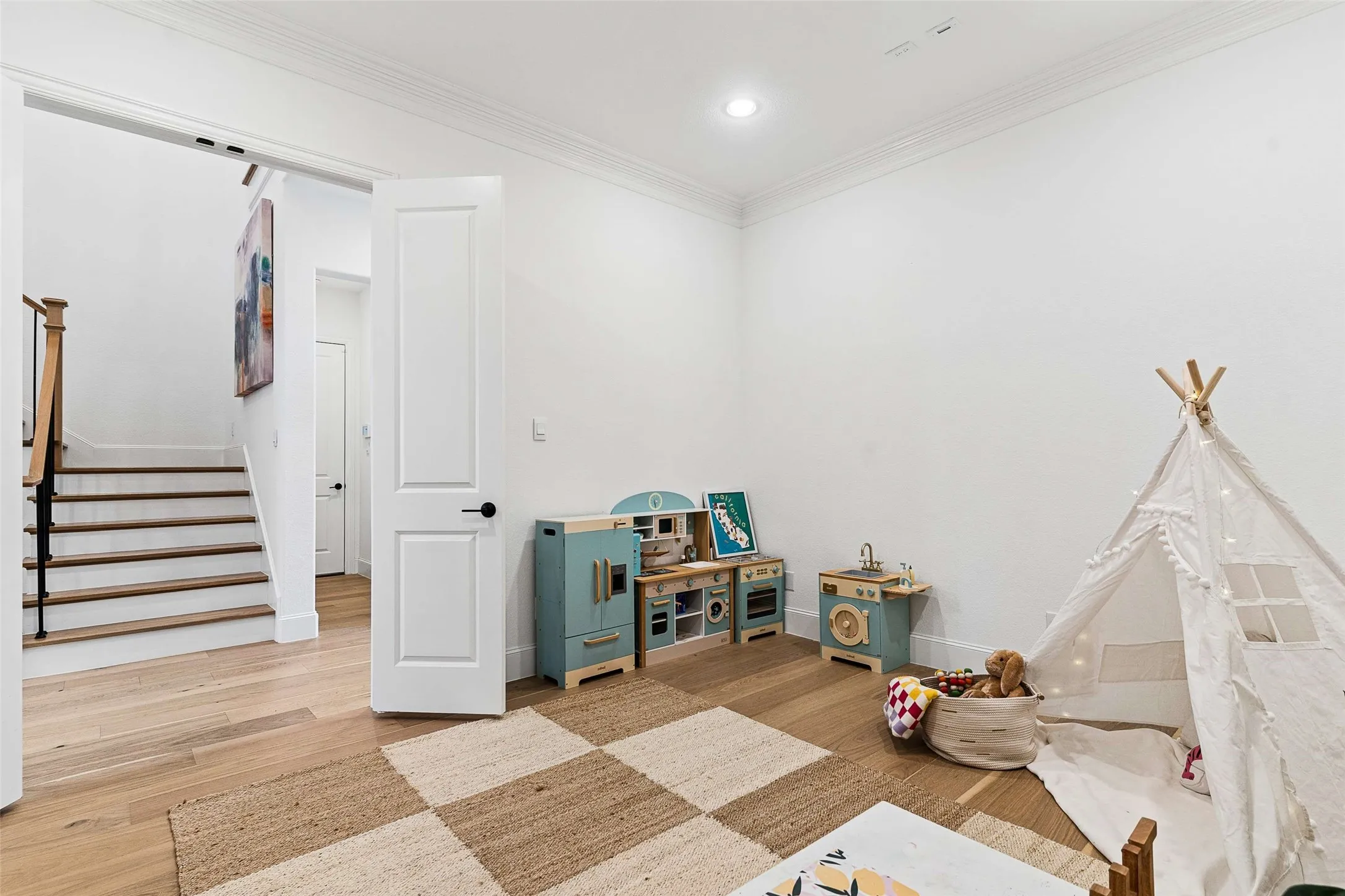 Bright room featuring wood-finish flooring, a white paneled door with matte black hardware, and a staircase with a wood handrail and black balusters