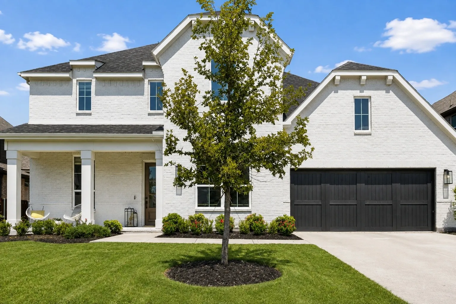 White brick exterior with a dark shingle roof, covered front porch, dark wood-finish front door, and a two-car garage