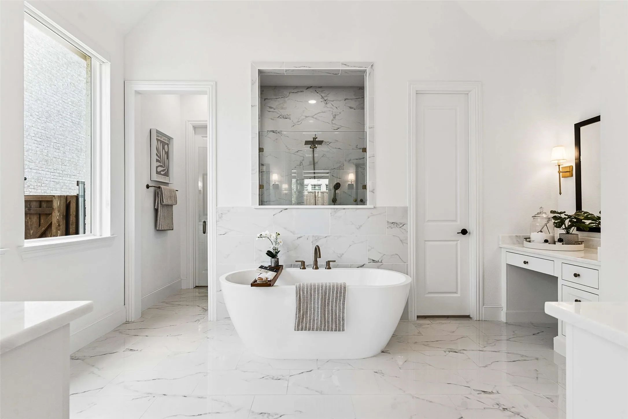 Freestanding soaking tub, walk-in shower with tile surround, vanity with built-in desk, large window, and marble-look floor and wall tile