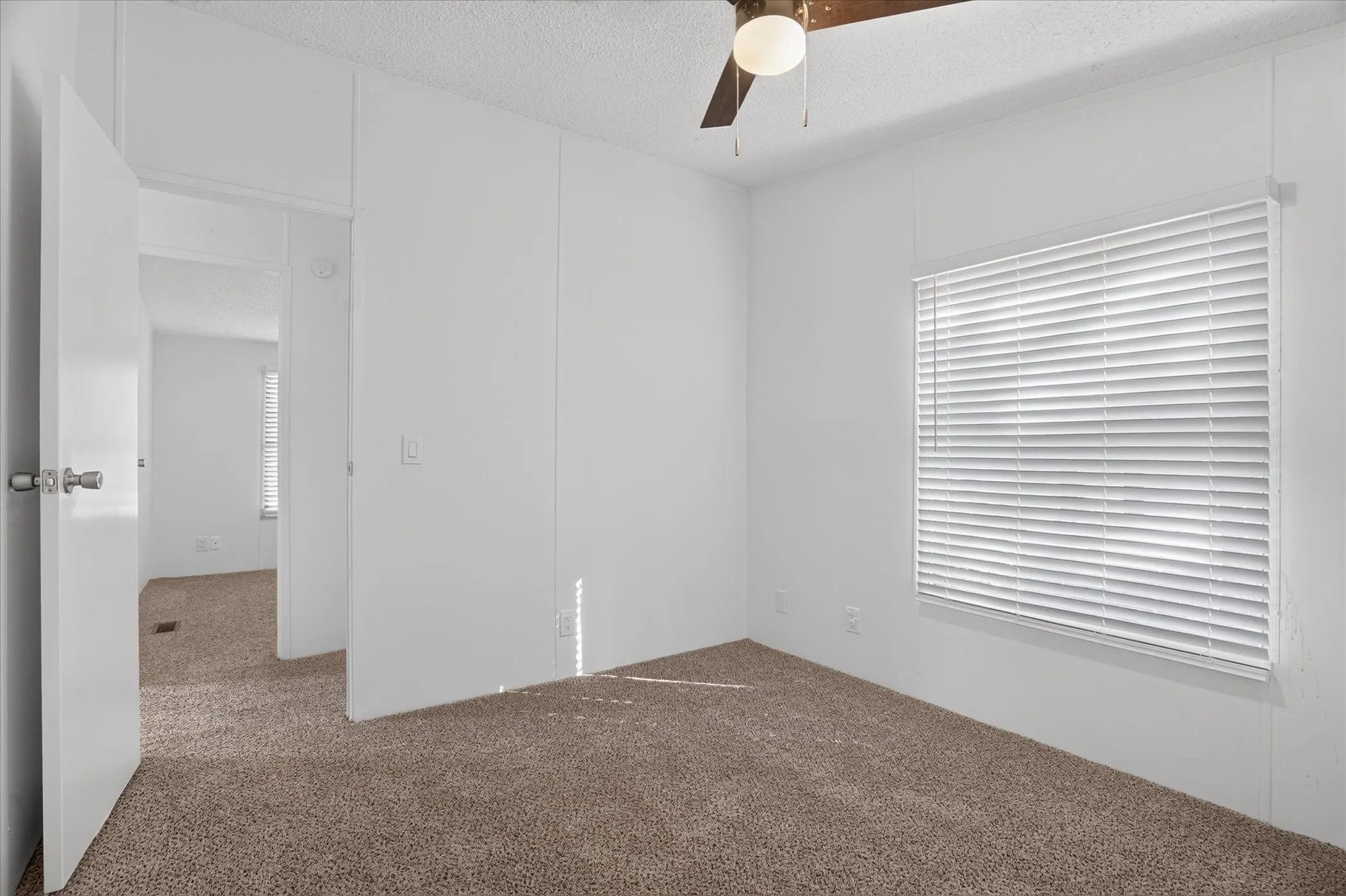 Neutral tone wall paneling, carpet flooring, window with blinds, ceiling fan with light fixture, and interior doors with lever handles