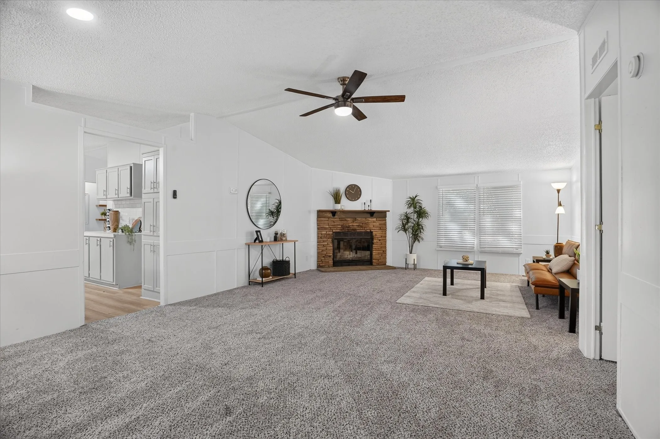 Spacious living area featuring a vaulted ceiling, brick fireplace, ceiling fan, carpet flooring, and a pass-through opening to the kitchen
