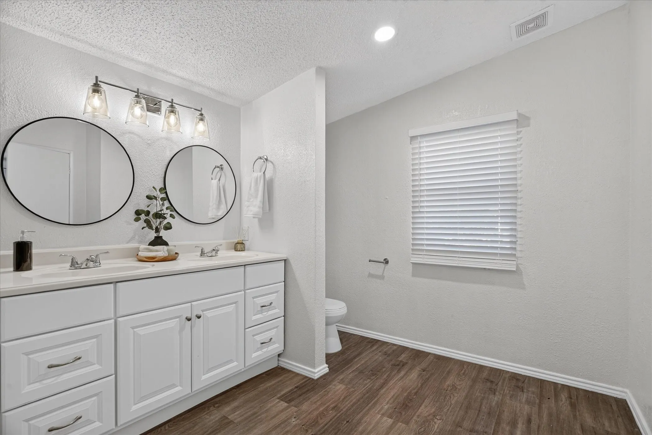 Dual vanity with white cabinetry and chrome hardware, individual round mirrors, a four-light fixture, wood-finish flooring, and a window with blinds