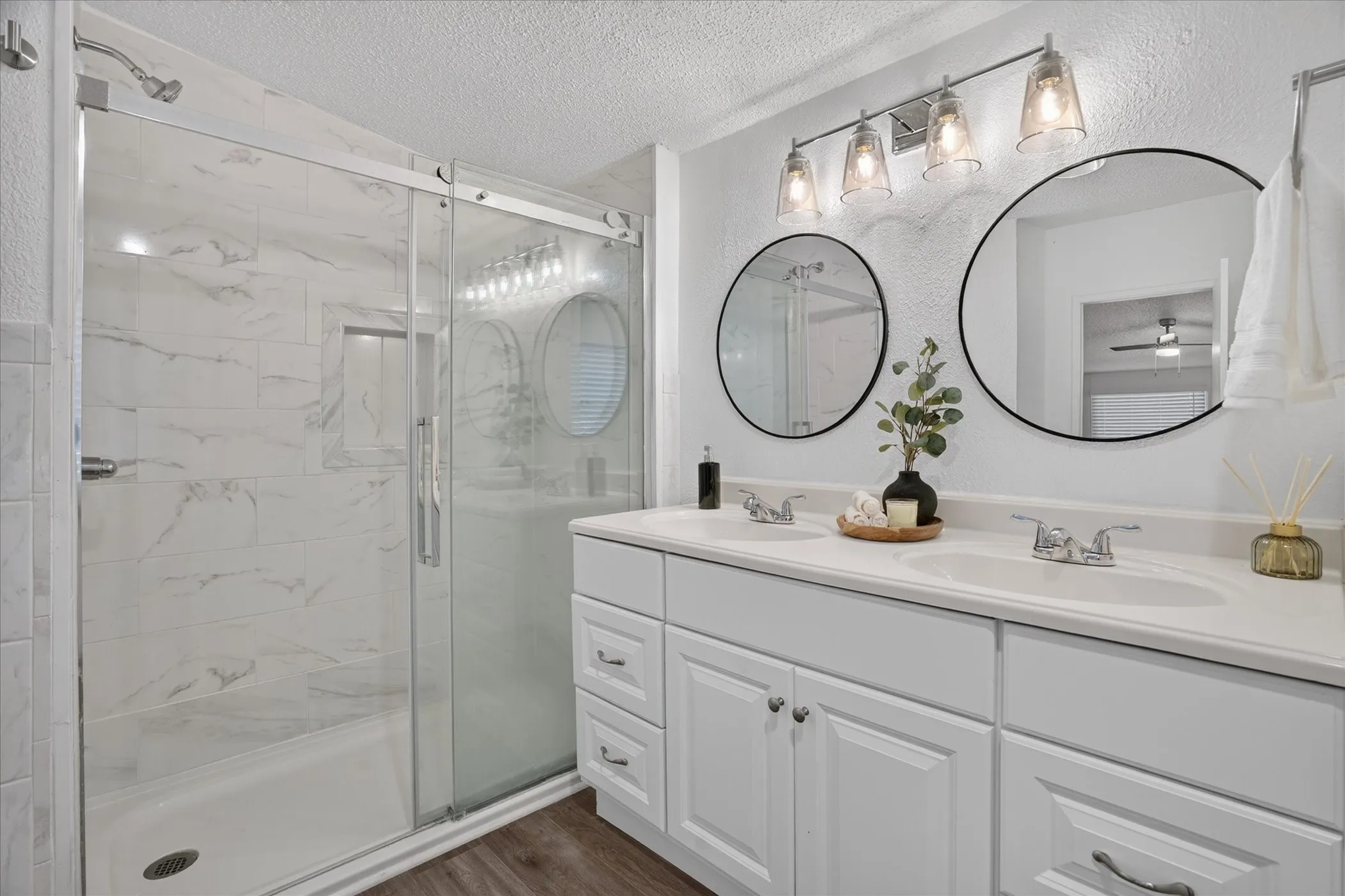 Dual vanity with white cabinetry, two round framed mirrors, and a glass-enclosed shower with white tiled walls featuring a built-in niche