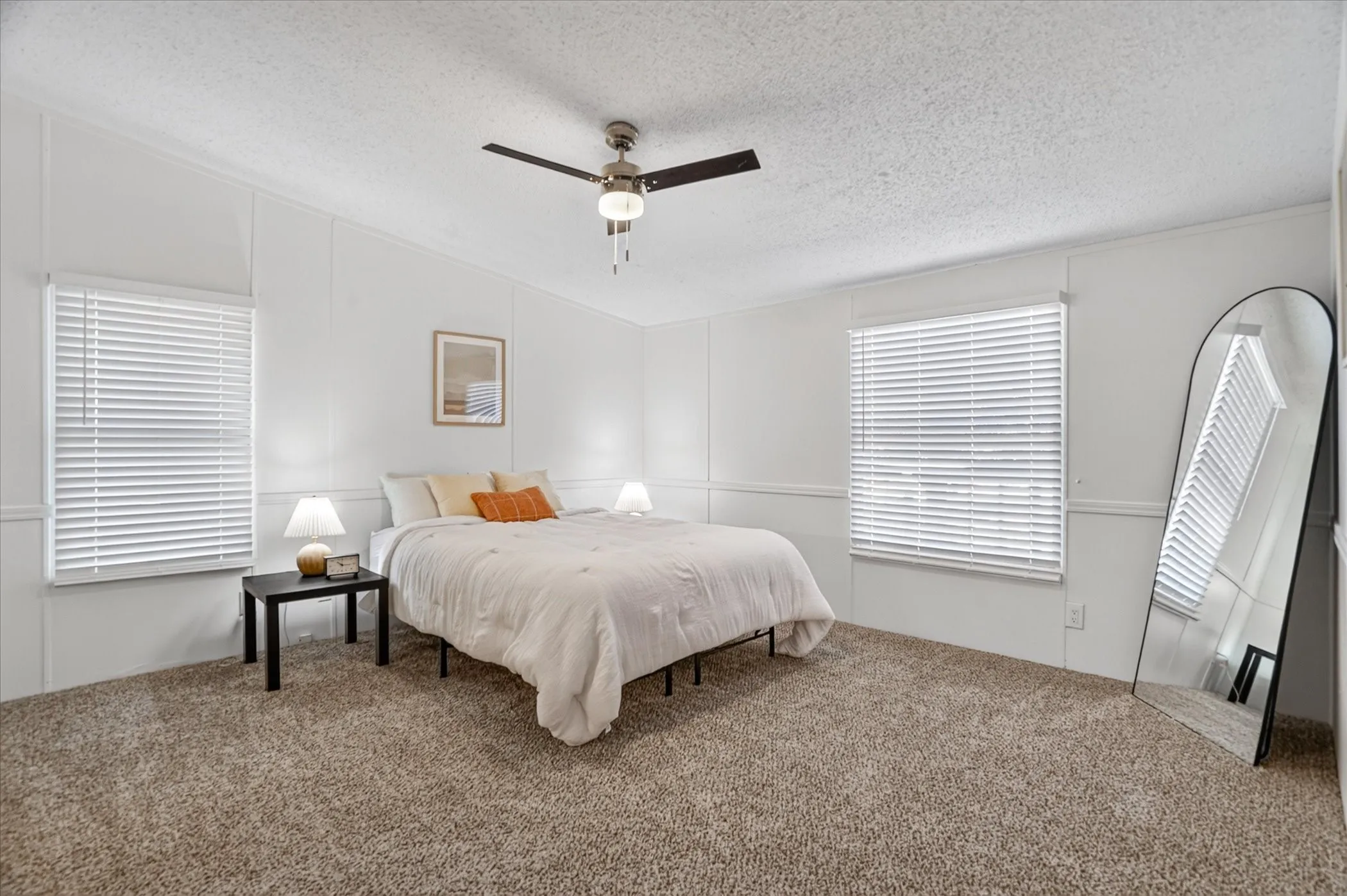 Spacious primary bedroom featuring neutral wall finishes, dual windows with plantation shutters, carpet flooring, ceiling fan with integrated lighting, and a full-length mirror