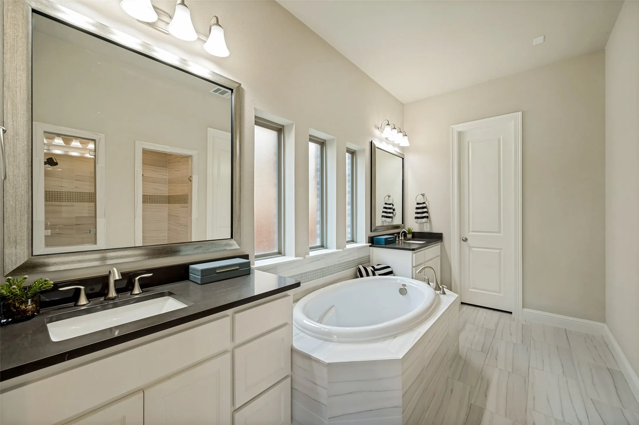 The luxurious primary bath is designed as a private spa retreat, featuring dual vanities, elegant framed mirrors, a soaking tub, and a beautifully finished walk-in shower. Abundant natural light, timeless tilework, and a bright, airy layout create a serene space that feels both refined and restorative.